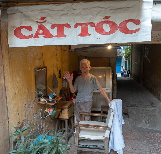 Picture of older man standing in outdoor alley, under a sign that says Cat Toc and next to a mirro hanging on the wall and a chairin front of the mirror. The man is waving to the camera.