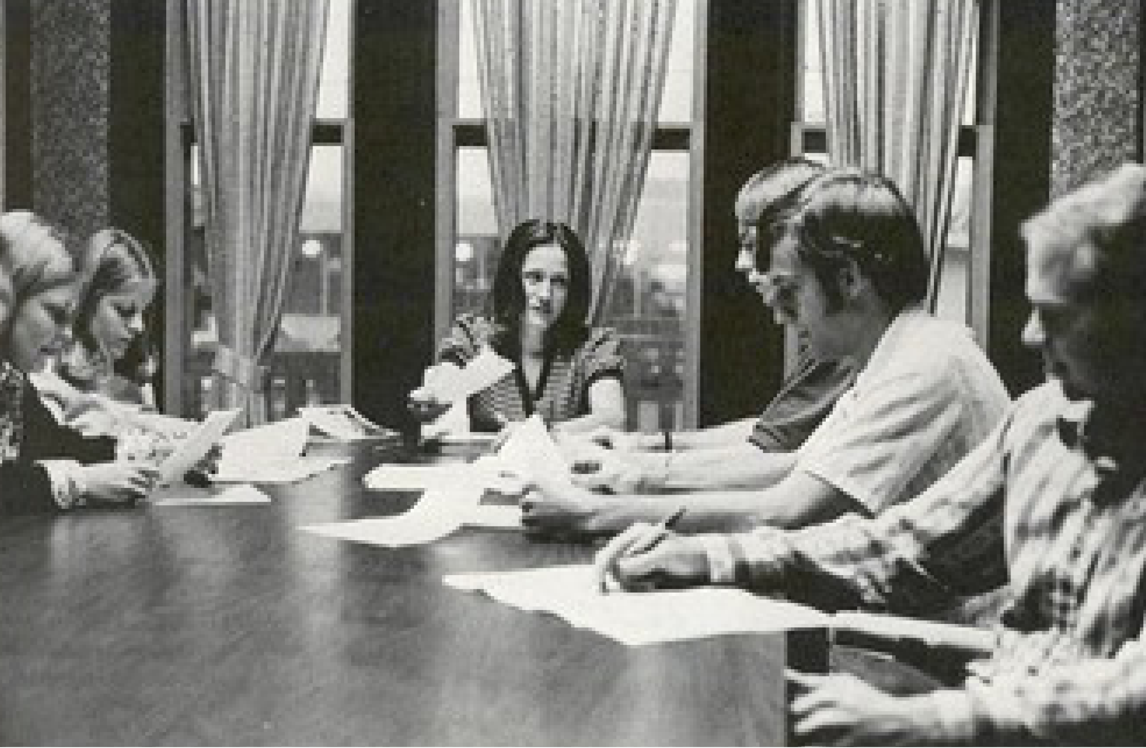 black and white photo of students sitting around a large table talking while holding papers