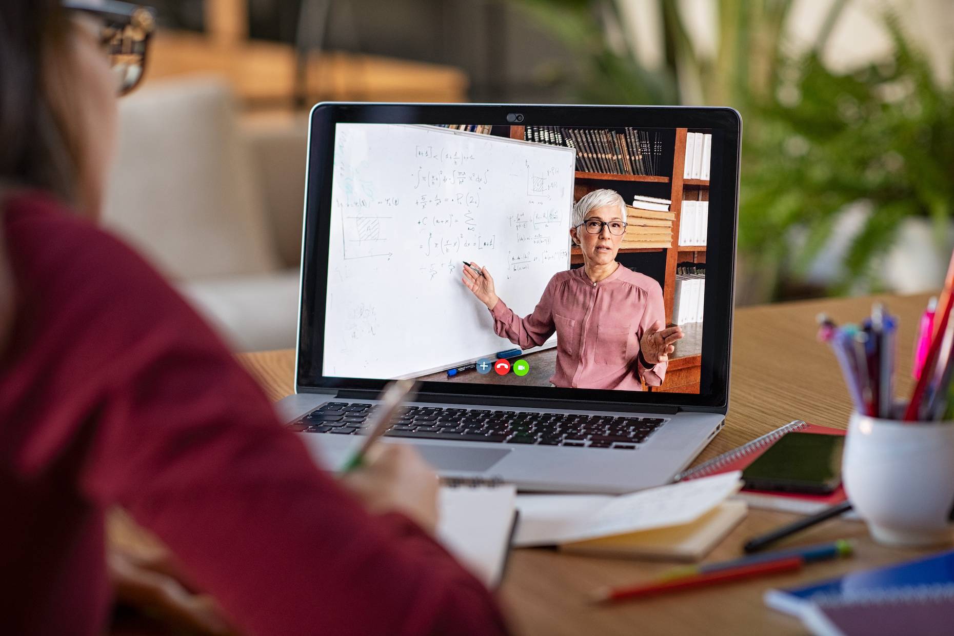Person watching a lecture on a laptop screen and taking notes. On the screen the instructor is pointing to a whiteboard with math equations