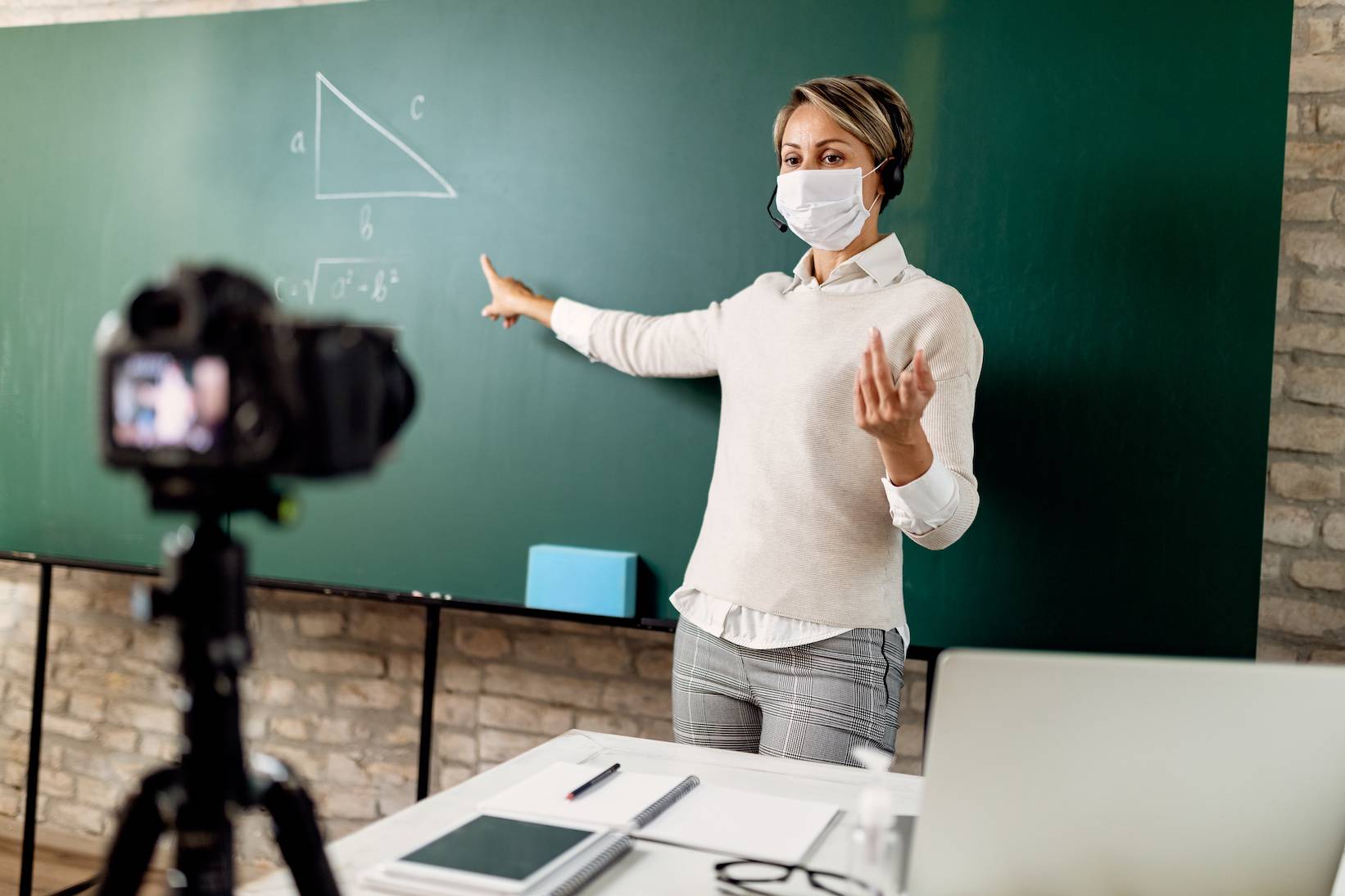 instructor giving a lecture in front of a chalkboard. A camera sits on the desk filming her