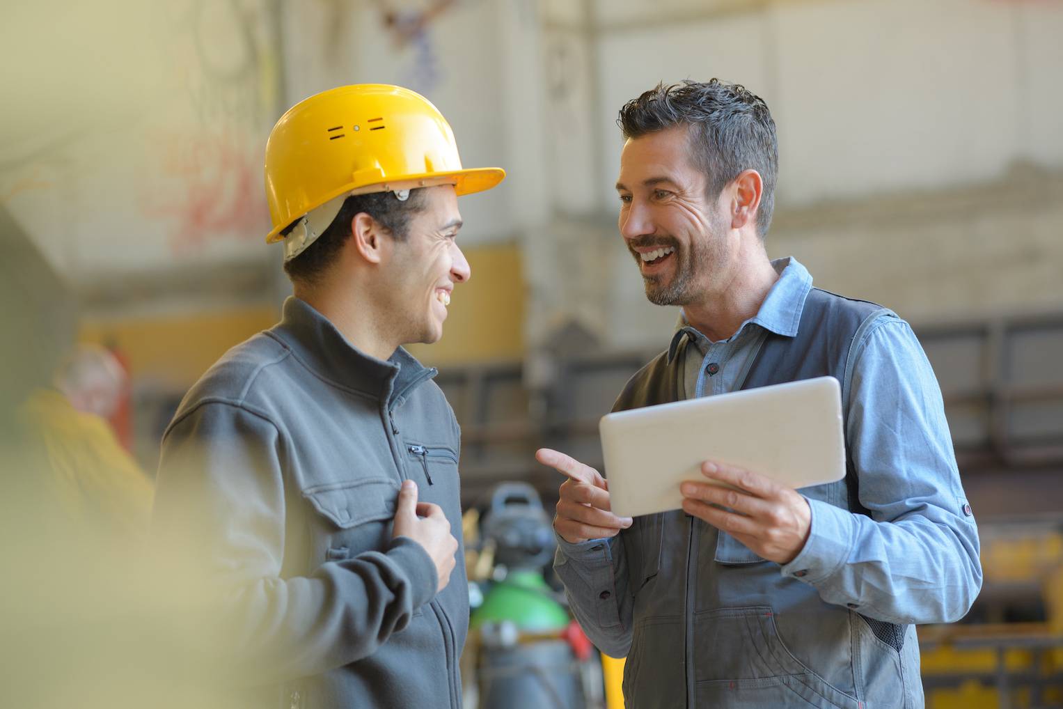 Two workers standing in a warehouse. The one on the left wears a yellow hard hat while the one on the right holds a tablet showing the screen to the other.