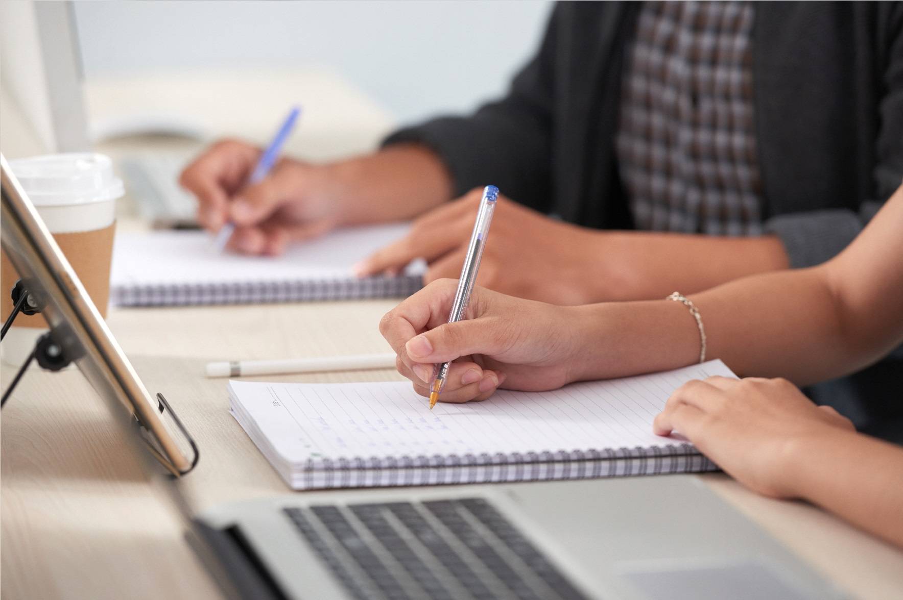 Laptops on a desk with hands writing on notebooks next to them.