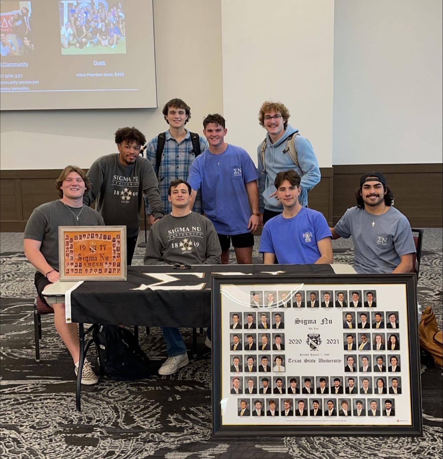 Eight men of Sigma Nu pose behind a table that features their organization's 1977 and more recent composite photo