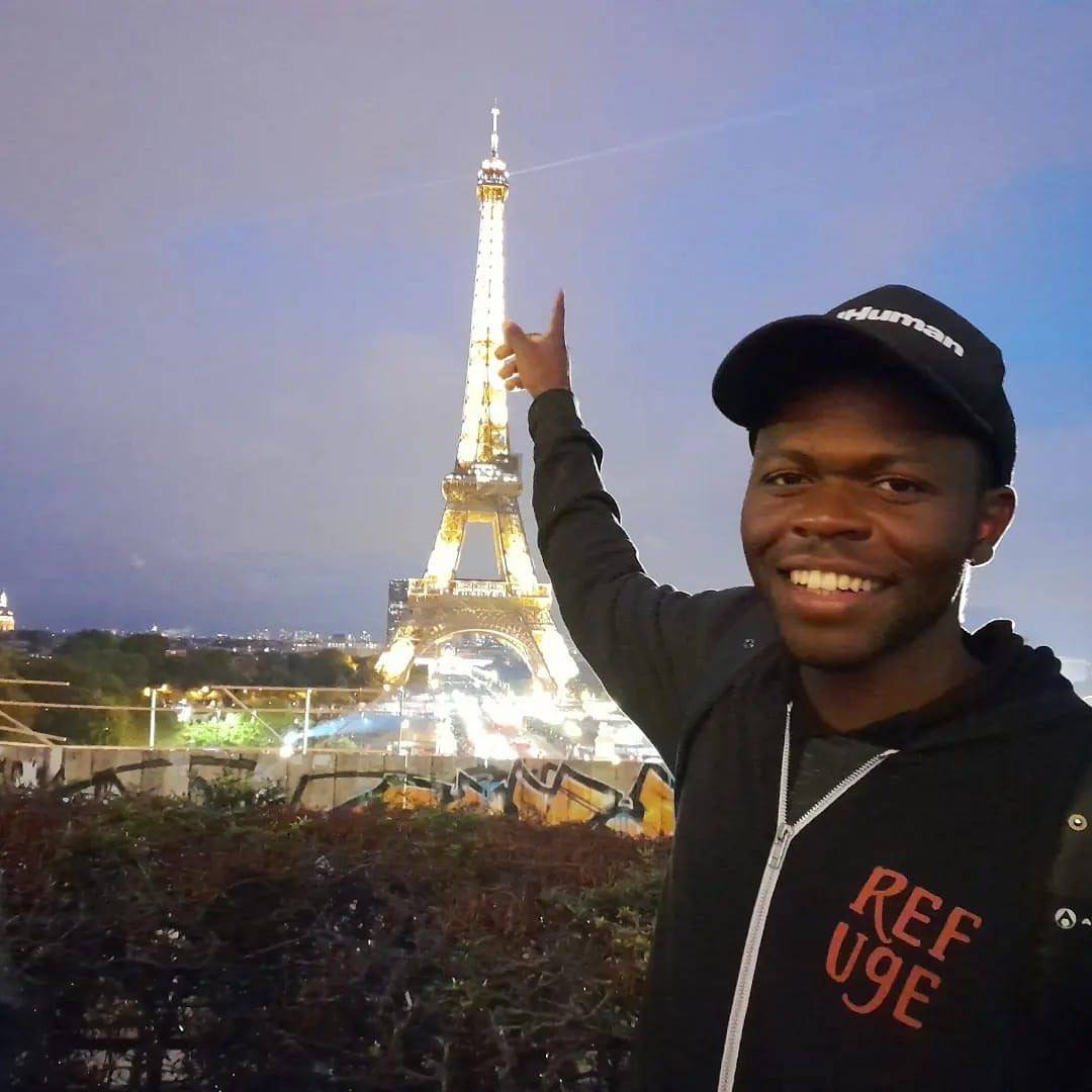 image: man wearing hoodie with "refuge" written on it, pointing at Eiffel Tower in background