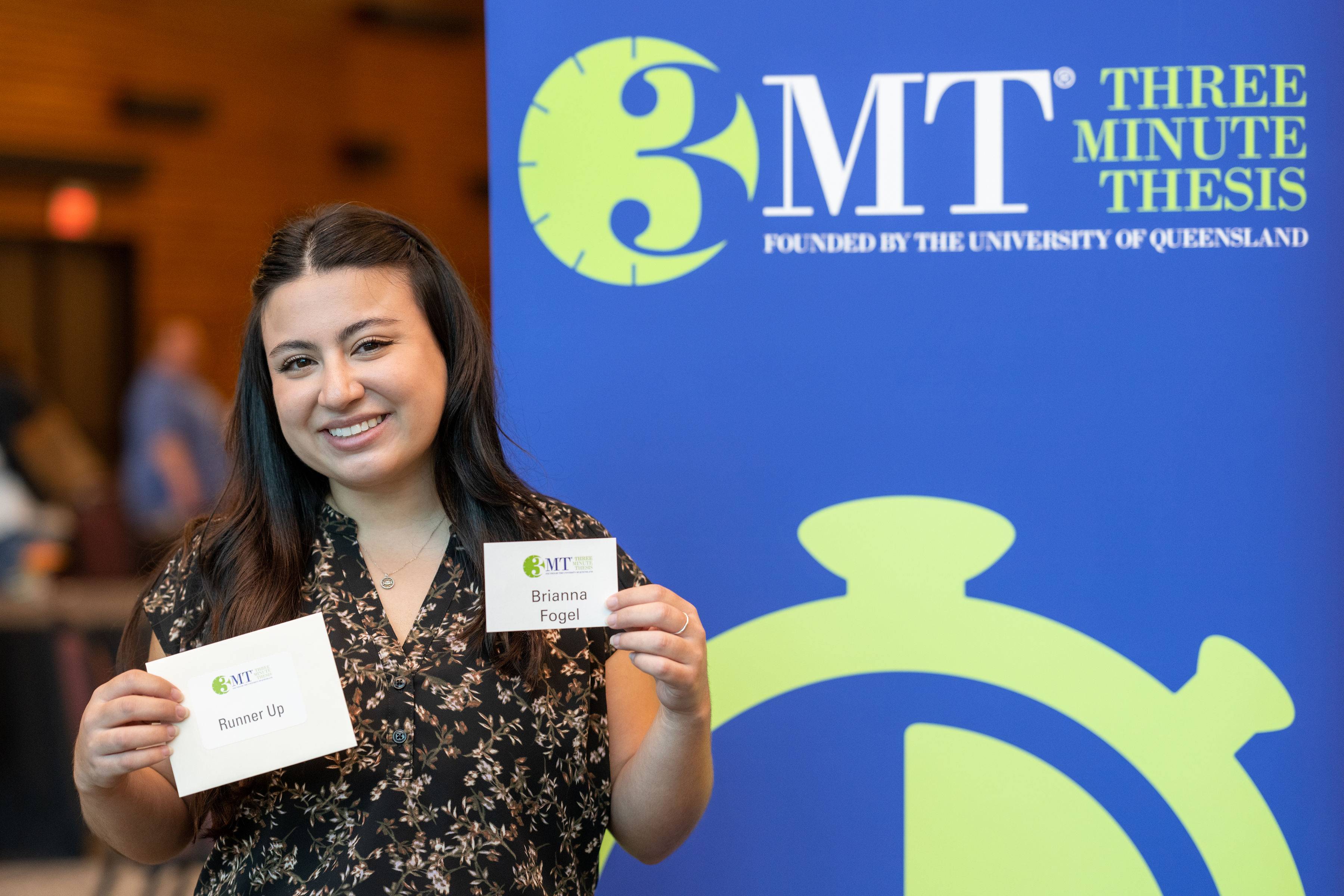Brianna Fogel standing next to a 3MT sign holding her name tag and runner-up annoucement