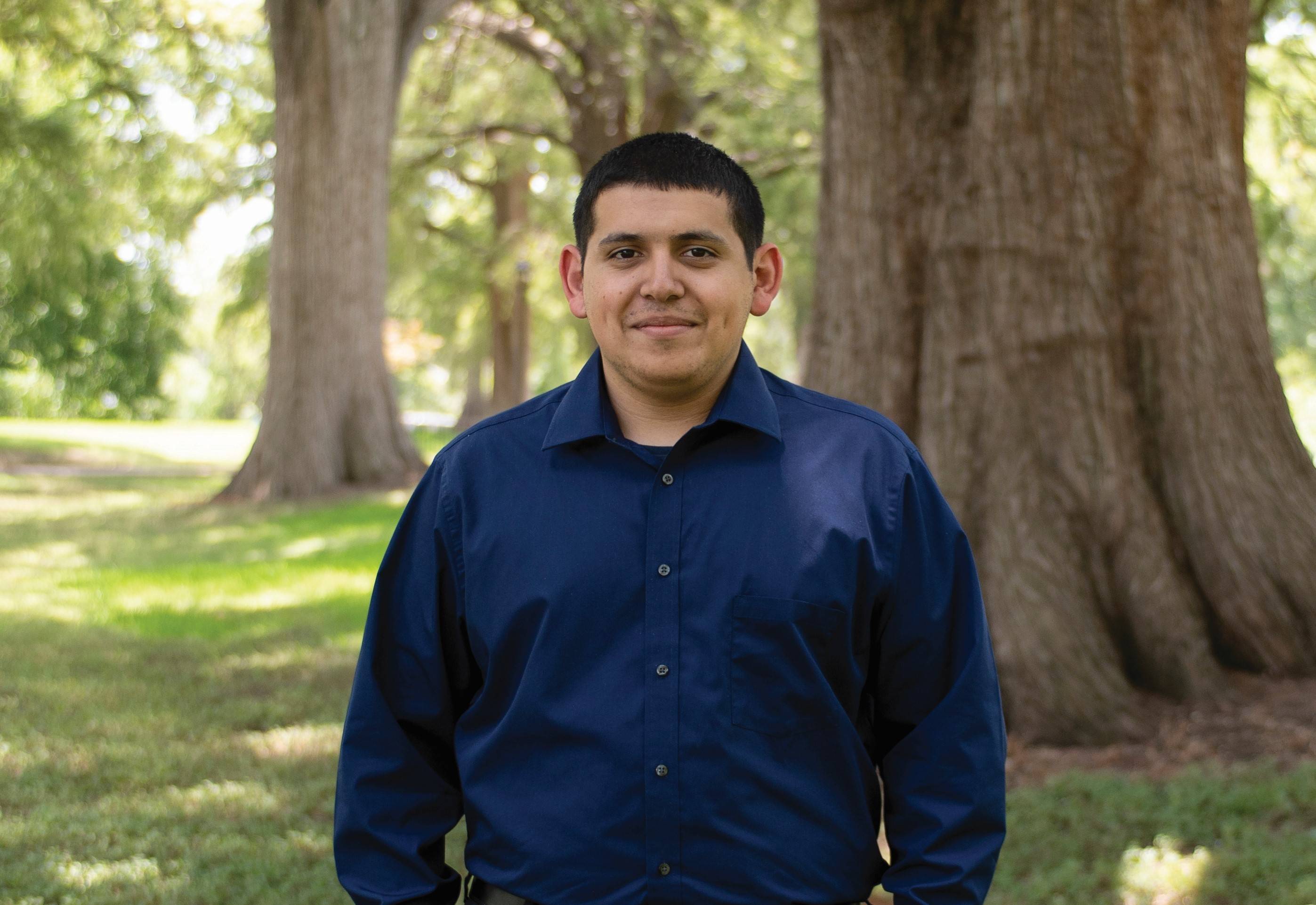 Christopher Solis smiling for a camera in a blue button up shirt in front of the ponds and large trees on east campus