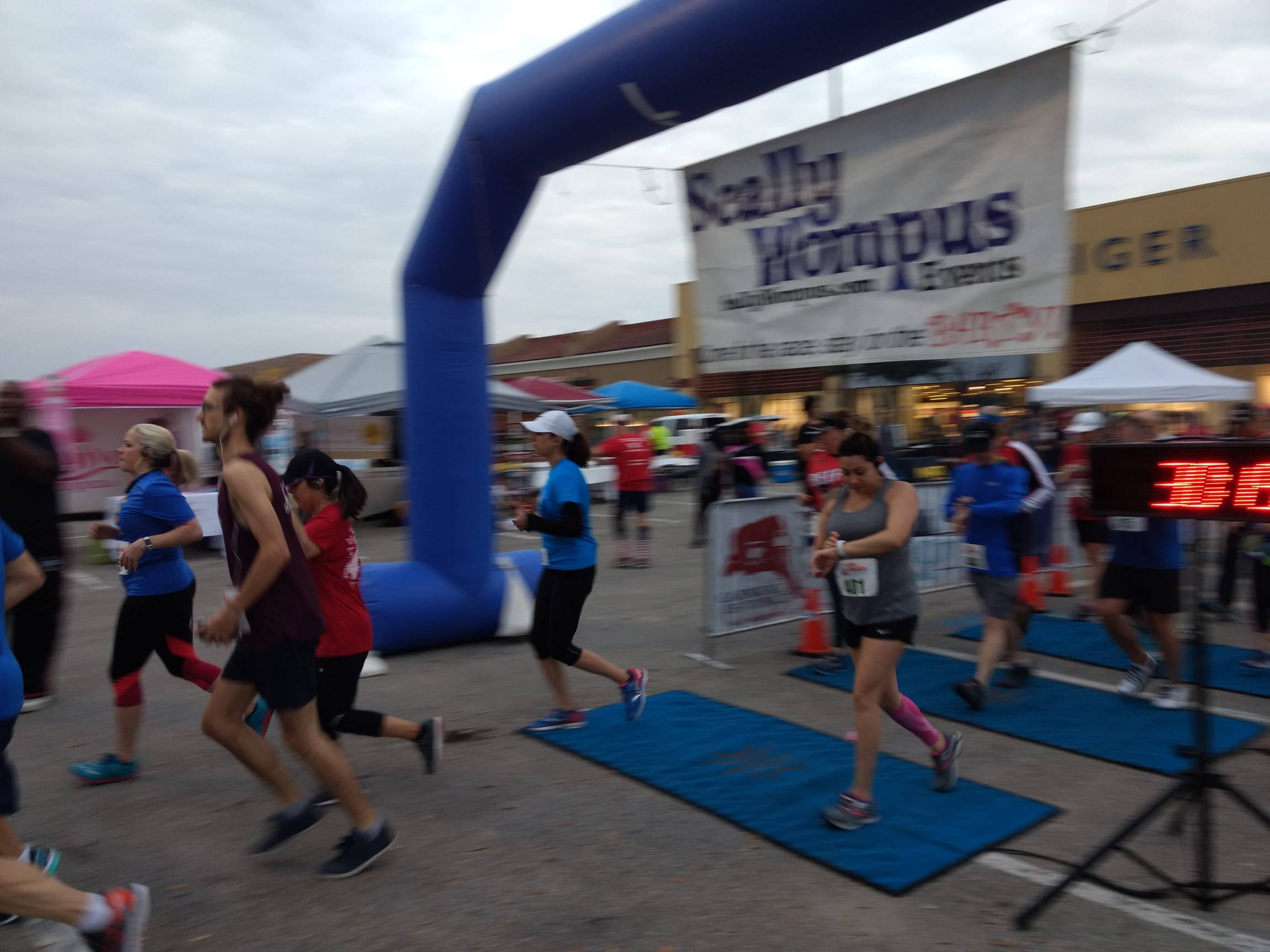A group of people running under a big blue banner at the start of the San Marcos Half-Marathon