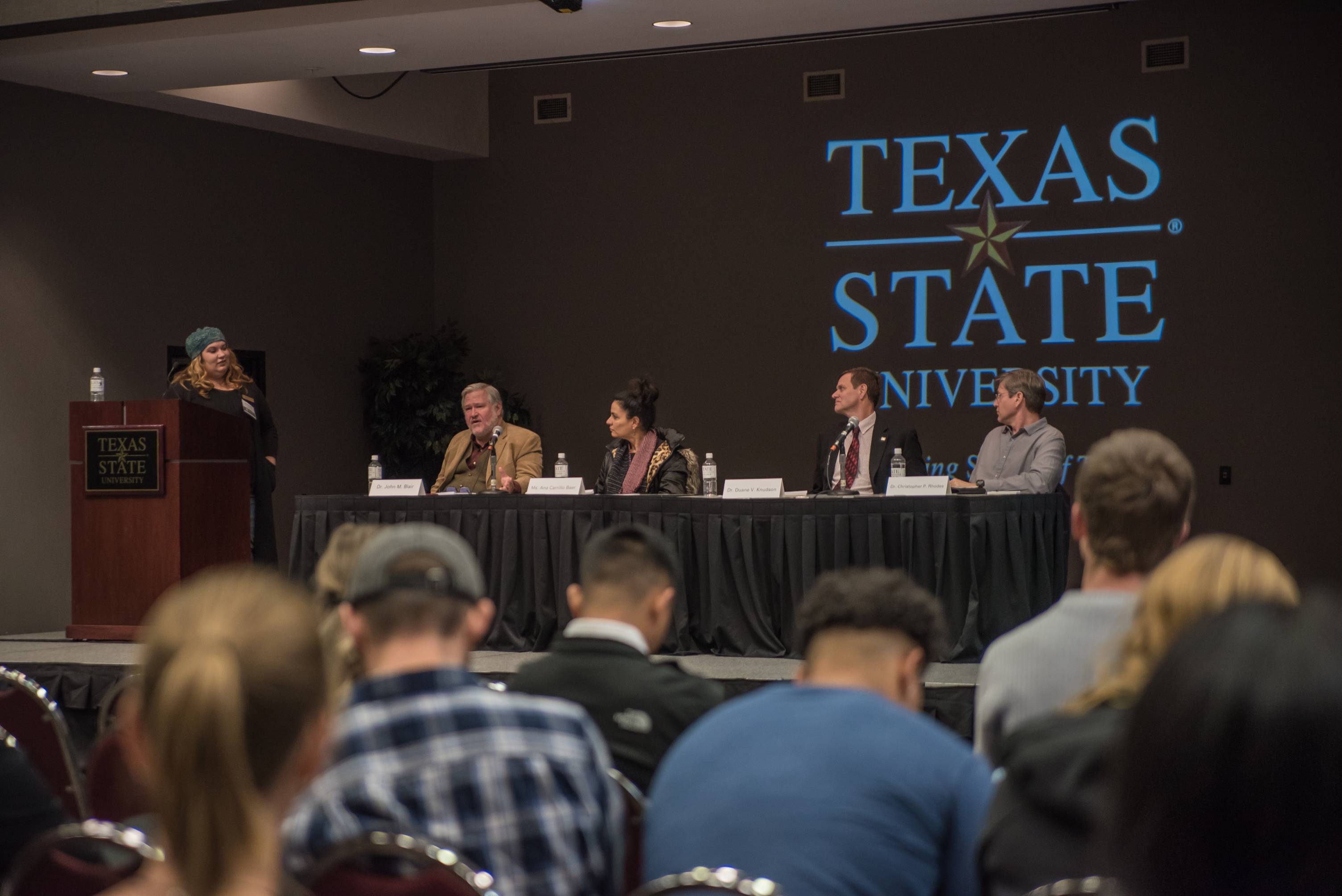 a panel of speaker sit at the front of a room in front of a large Texas State logo