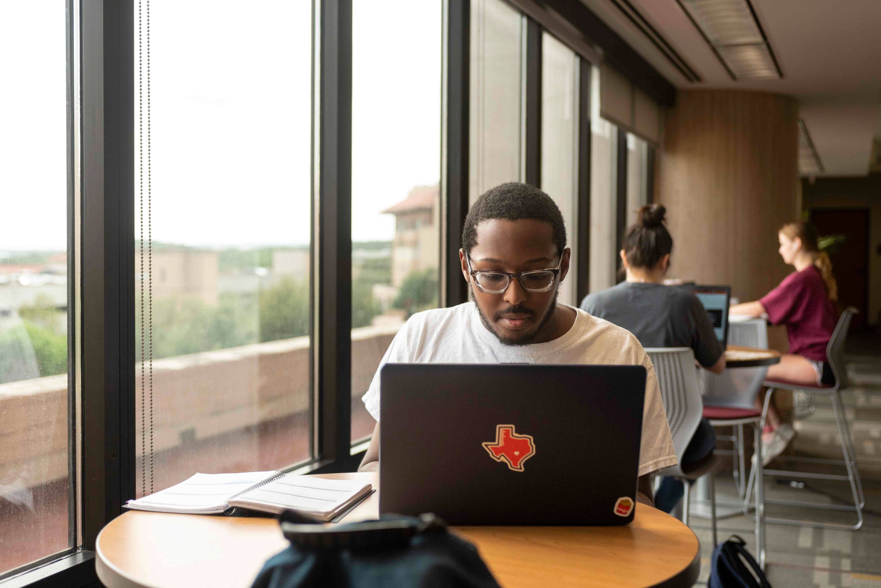 a black male student works diligently on his laptop by a window inside the Alkek Library