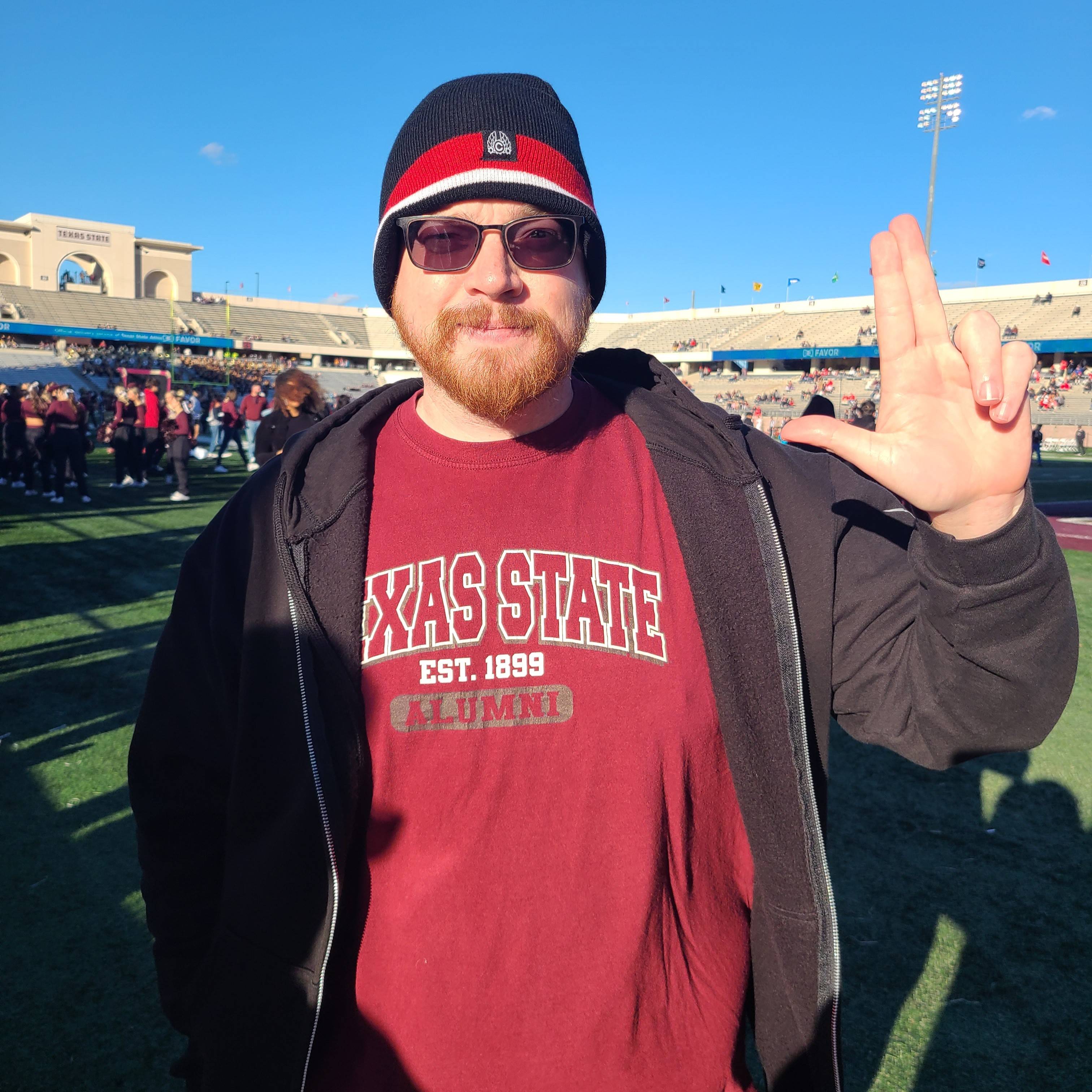 Mark Moore wearing a maroon TXST t-shirt, with a black hoodie, and a black and red beanie while holding up the TXST hand symbol with his left hand on a football field