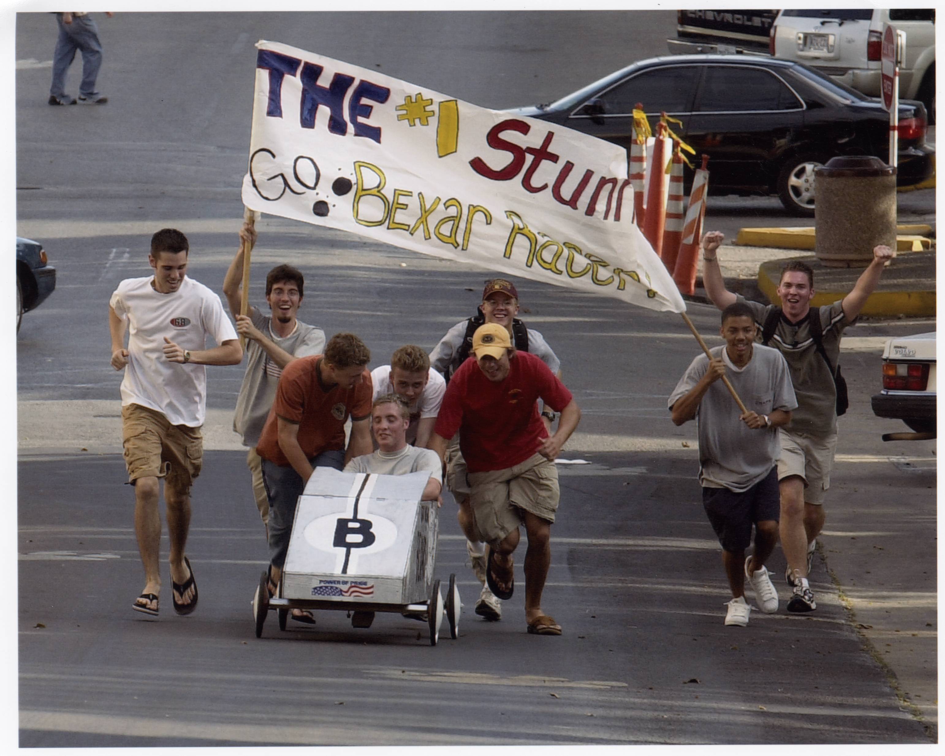 photo from 2002 of men pushing soap box car down street while holding up large flag