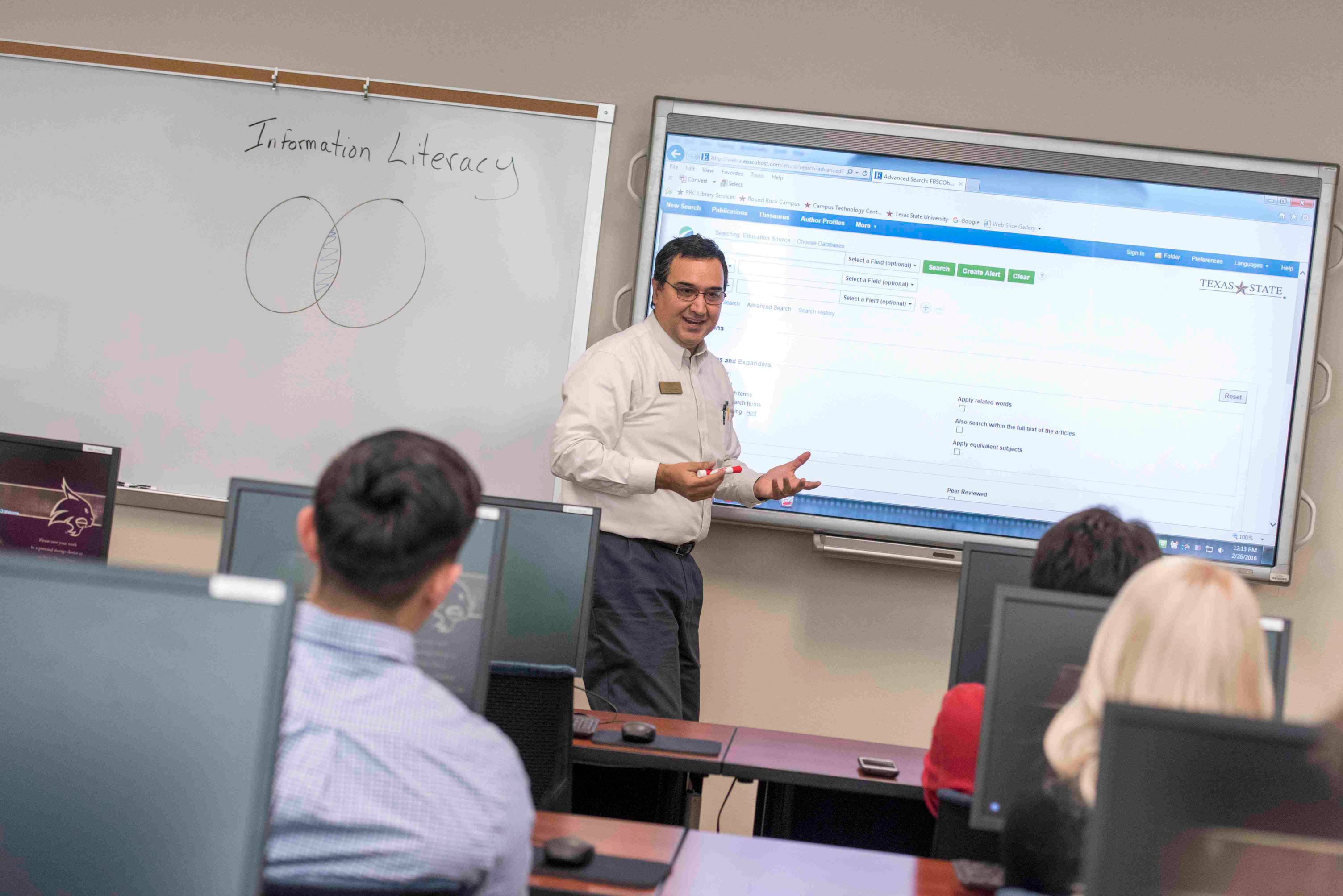 a male professor stands in front a projected computer screen as he lectures a small classroom