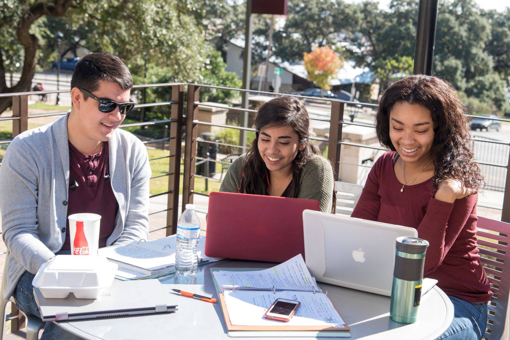 three students sitting at a table outside with laptops