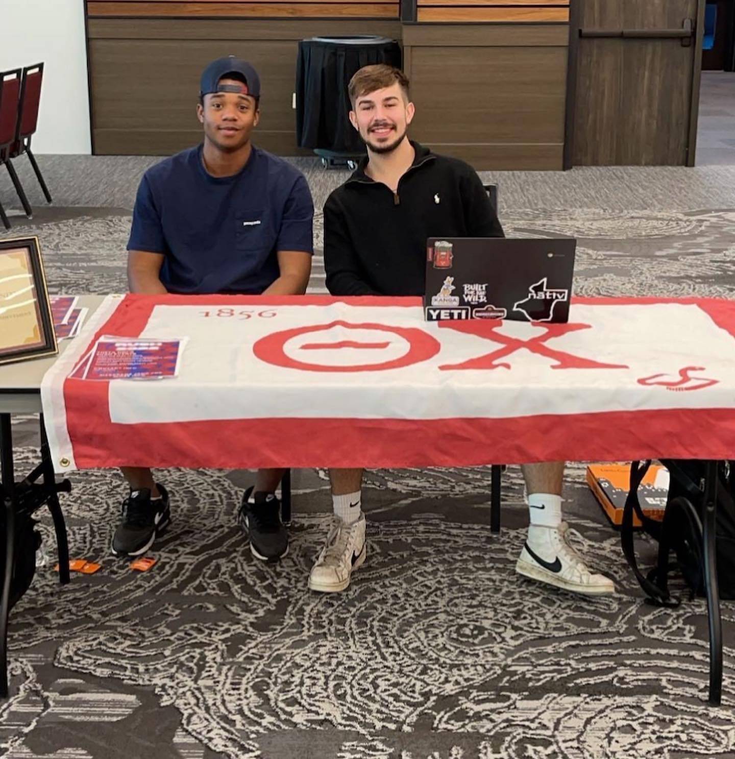 Two Theta Chi men sitting at a table with their flag draped over the top