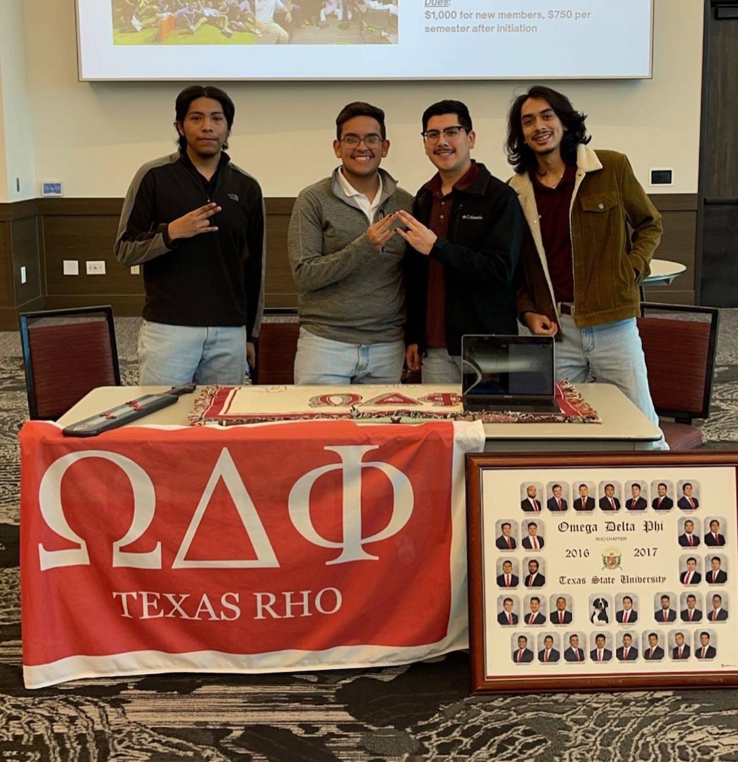 Four men of Omega Delta Phi posing at a table featuring their organization's flag and composite photo