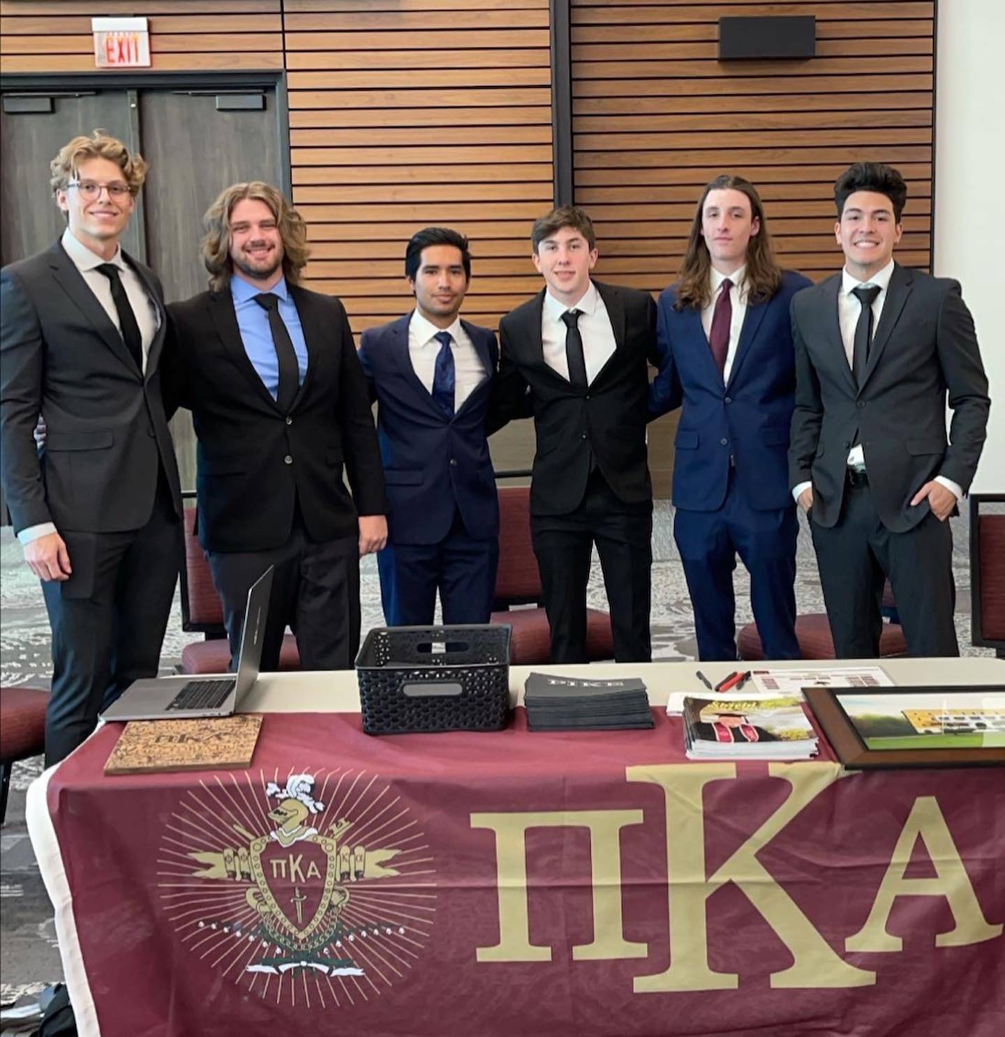 Six men of Pi Kappa Alpha stand behind their table/organization banner wearing suits with ties