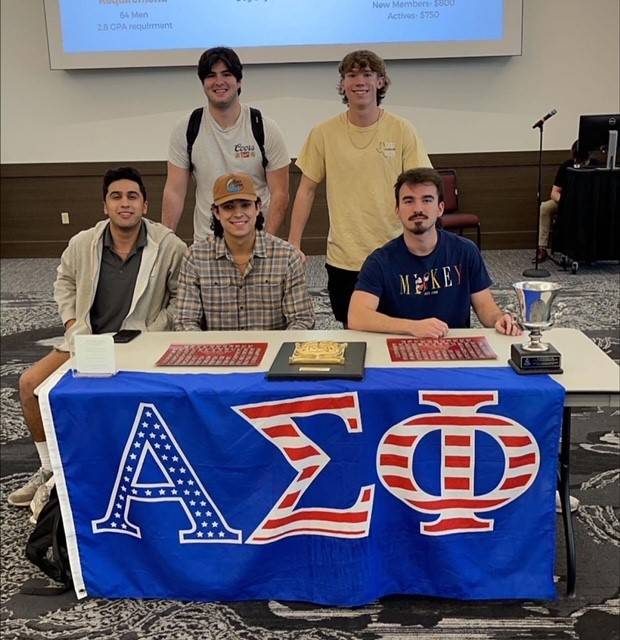 Five members of Alpha Sigma Phi post behind a table with their Greek Letters on the tablecloth