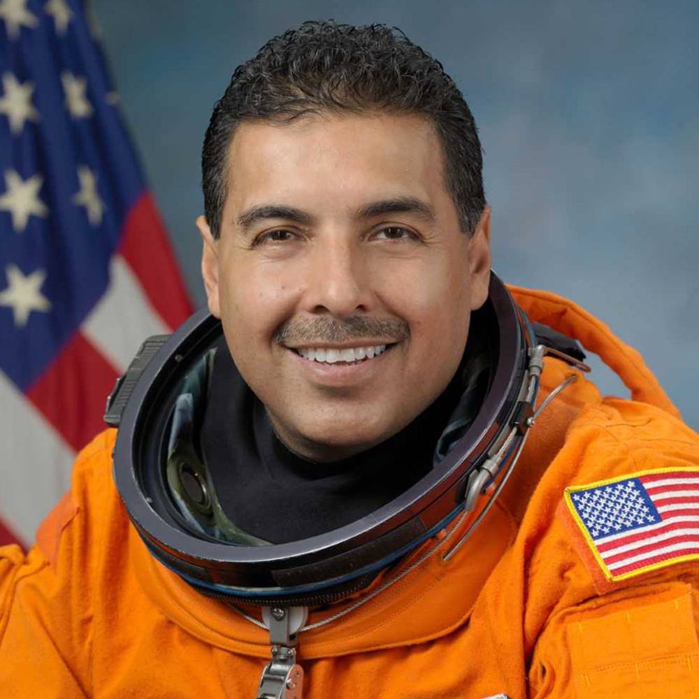 Smiling man wears an orange astronaut suit, the American flag seen in the background