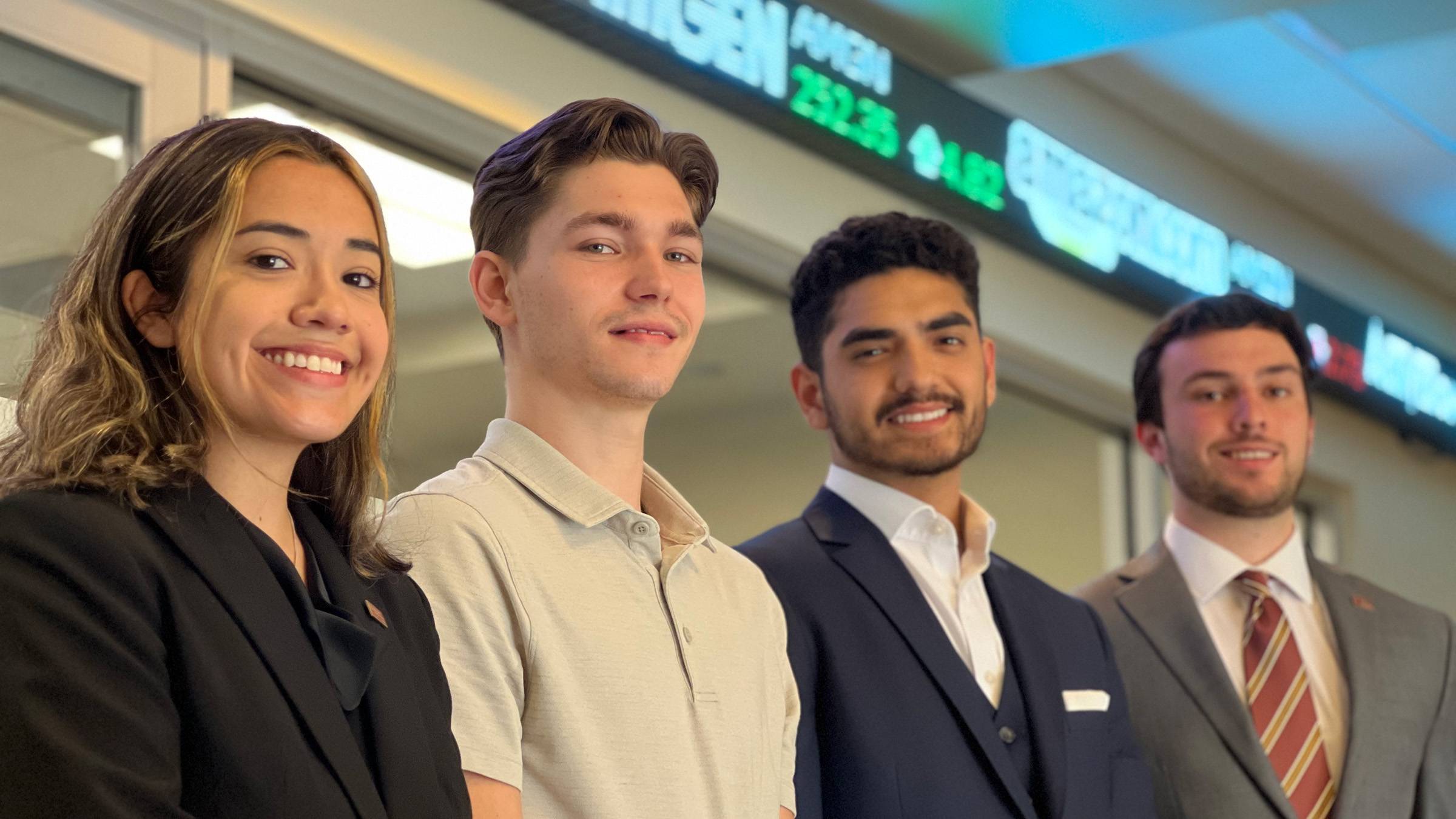 Four students in business attire pose in front of digital stock ticker