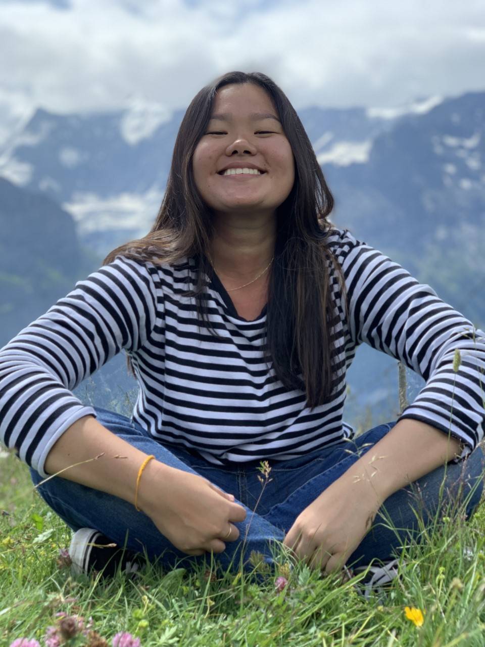 young woman sitting cross-legged in grass, smiling, with mountains and sky in background
