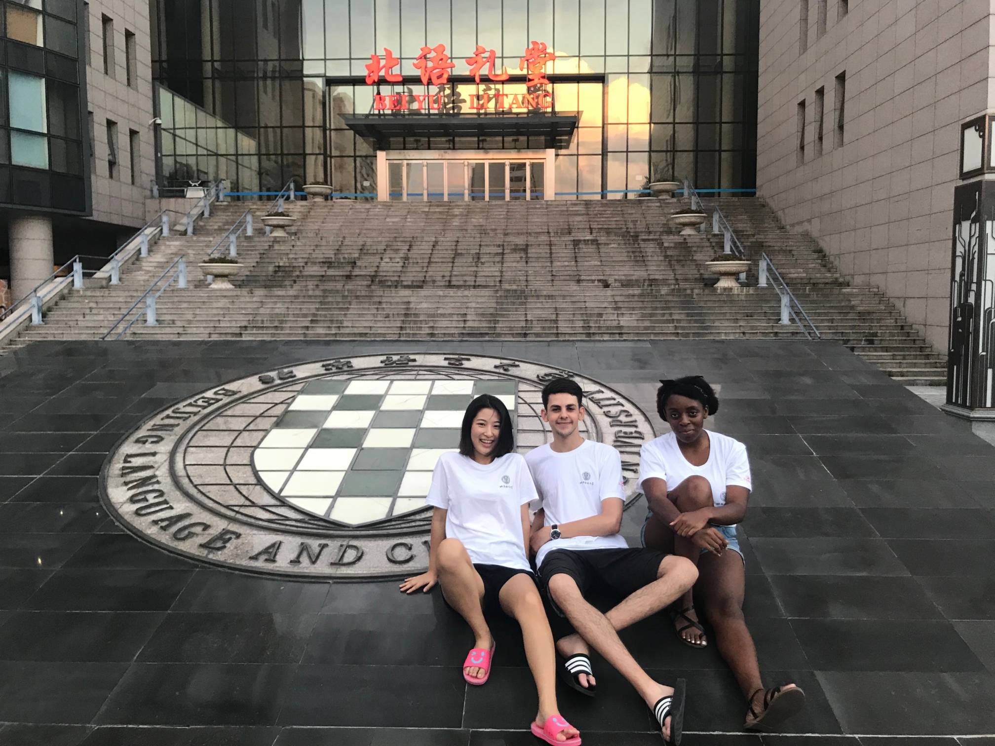 two women, one man sitting on ground in front of building, text embedded around seal reads "Beijing Language and Culture University"