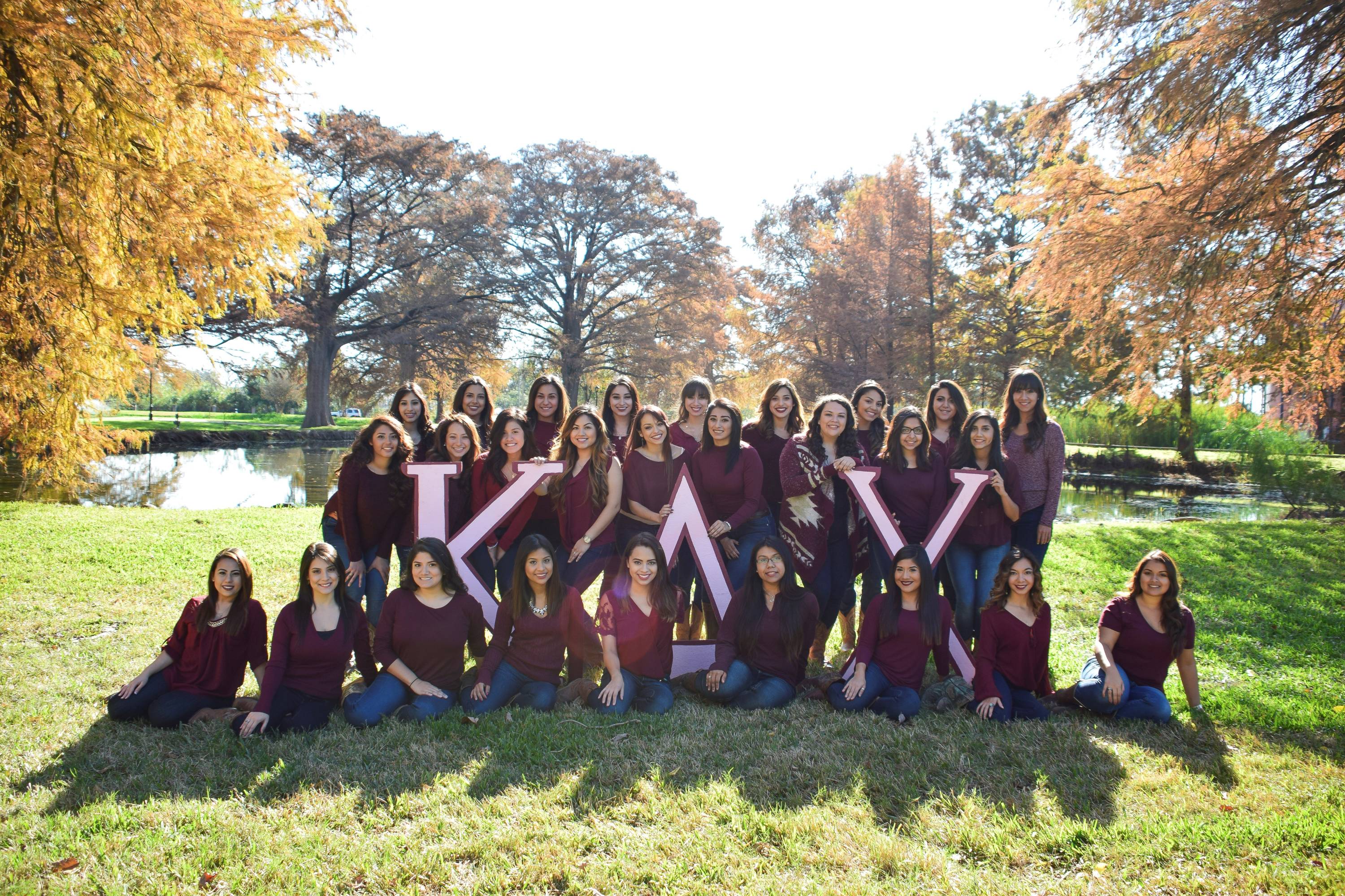 Women of Kappa Delta Chi all matching wearing jeans and maroon tops, posing in front of the ponds by the Theatre Building