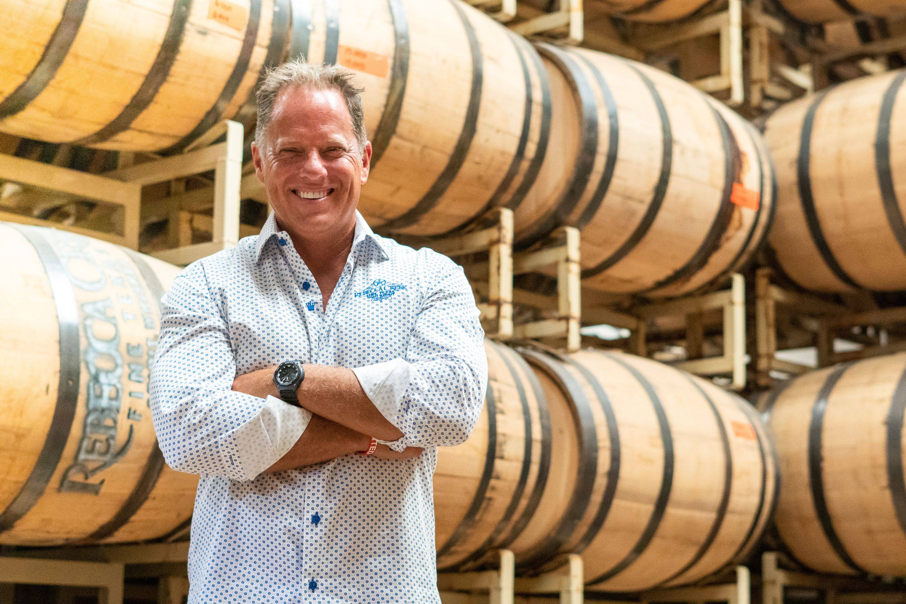 man smiling in front of wine barrels with arms crossed