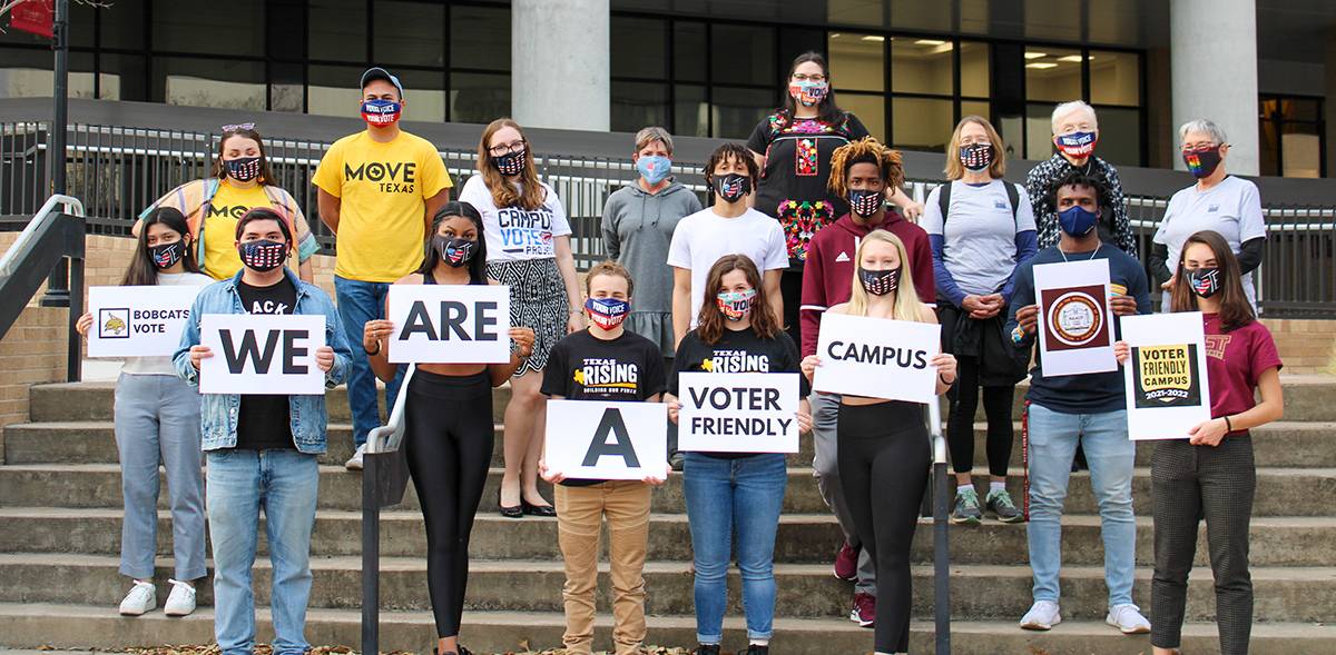 group of people standing on steps holding signs reading "we are a voter friendly campus"