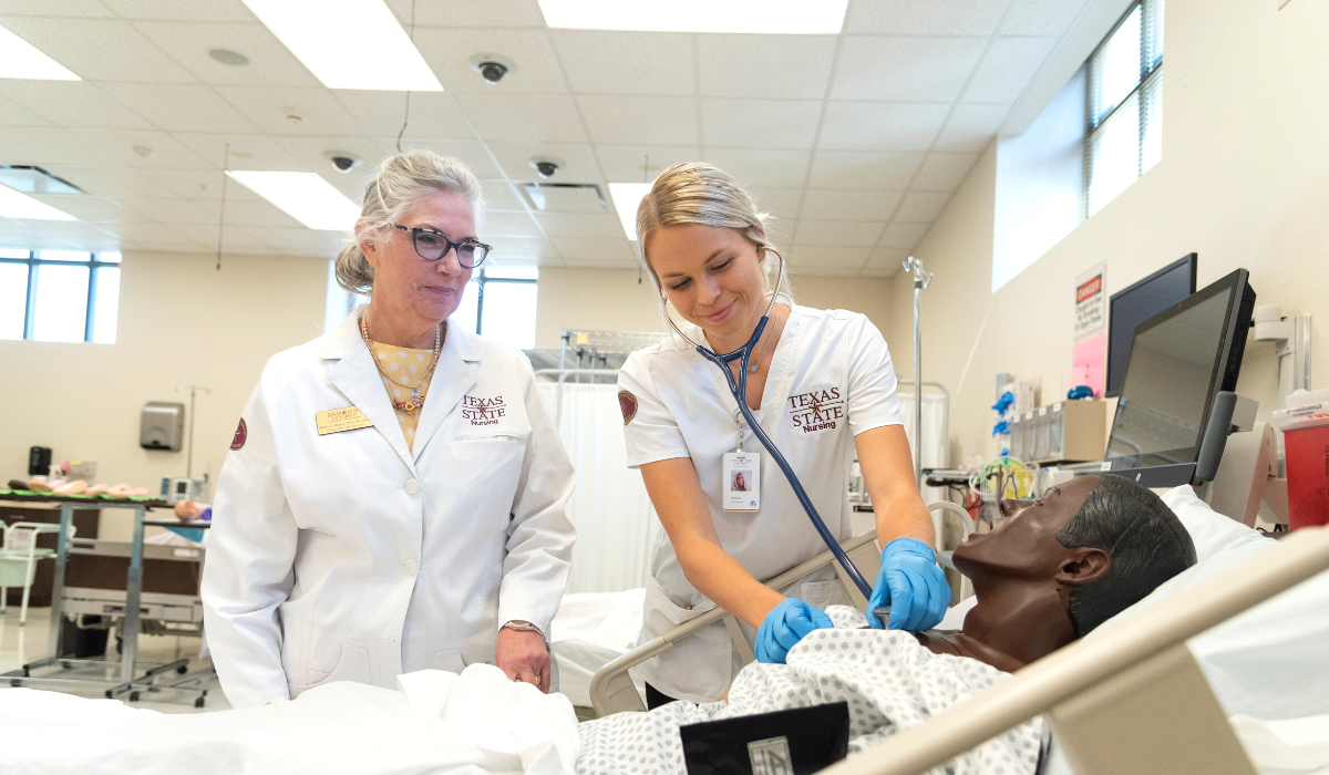 A female nurse checking the heart rate of a dummy patient while a older female watches her to her right.