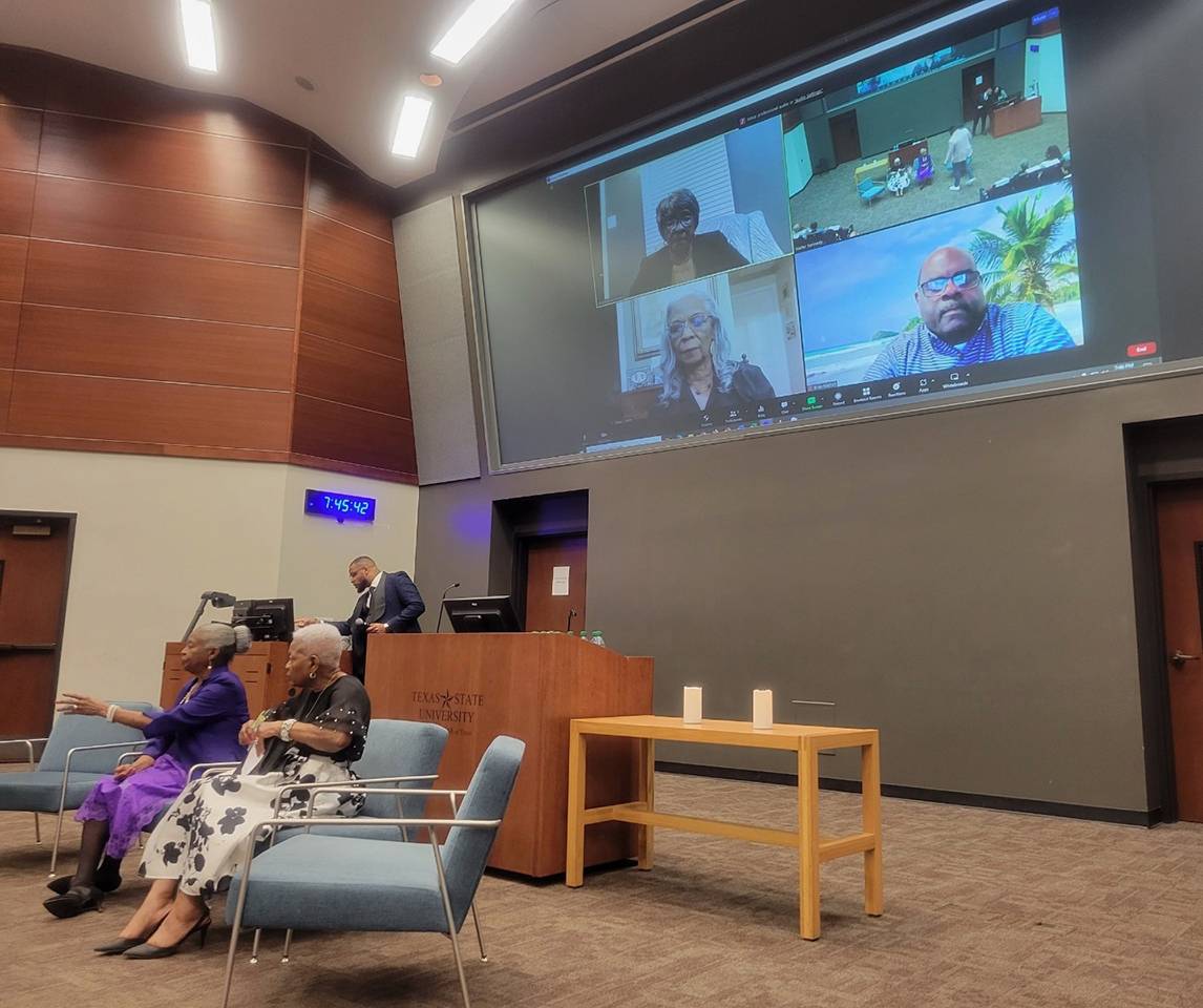 large screen with people on video call above two women sitting in chairs