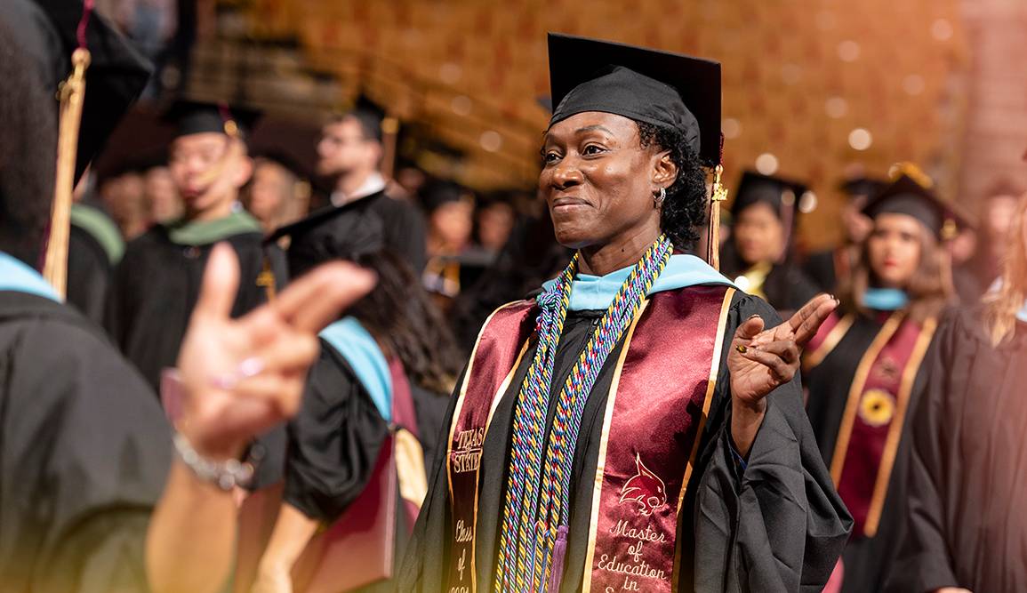 female graduate in cap and gown holding txst hand sign