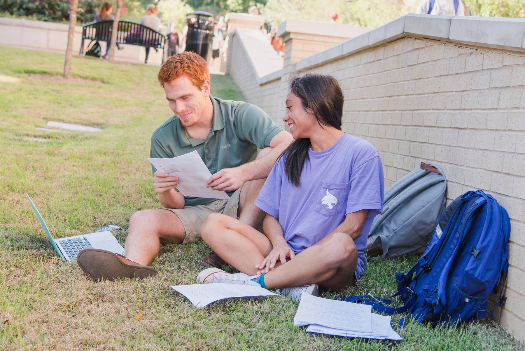 two students sitting outside and looking at a paper together. their backpacks are next to them.