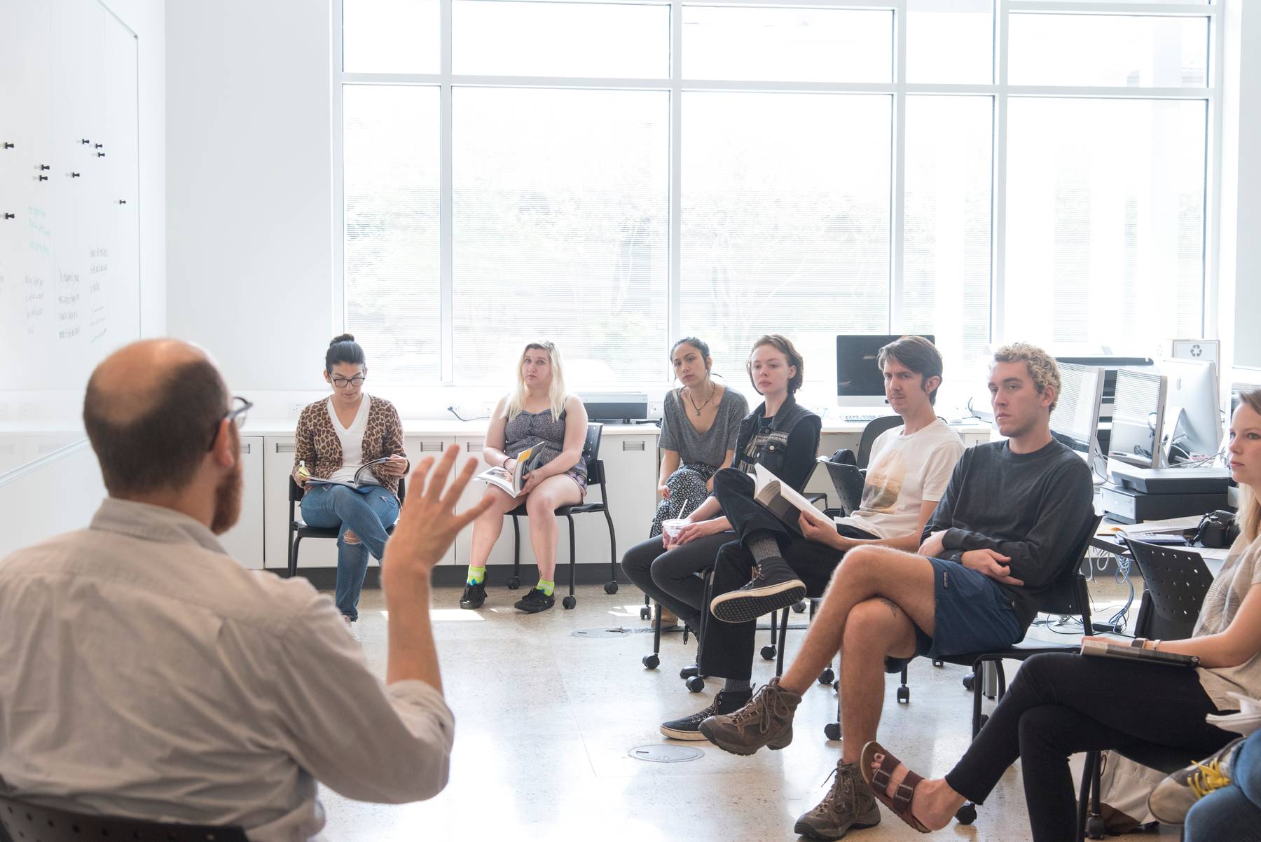 Students sitting in chairs listening to lecture surrounding the male lecturer in a bright classroom setting.