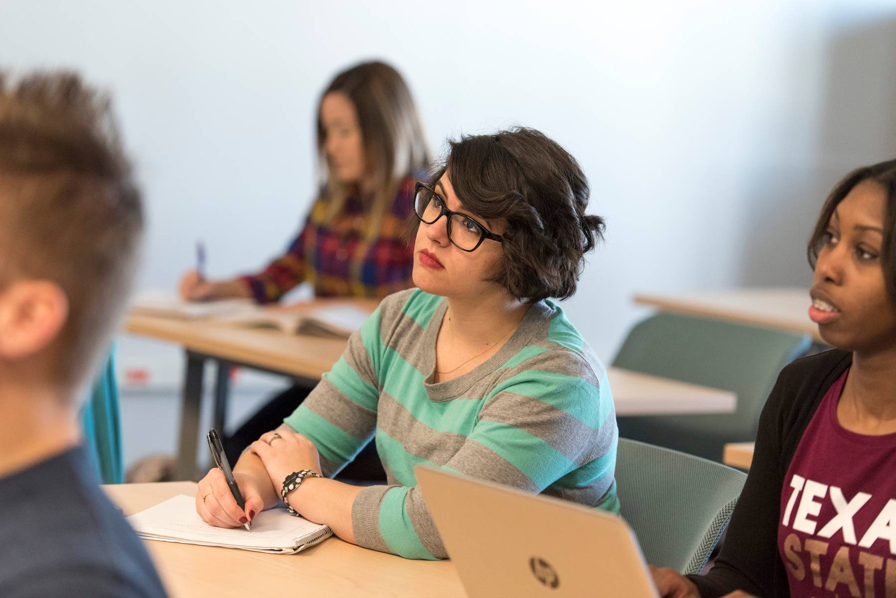 a student sits at a desk alongside other students as she diligently takes notes during a presentation