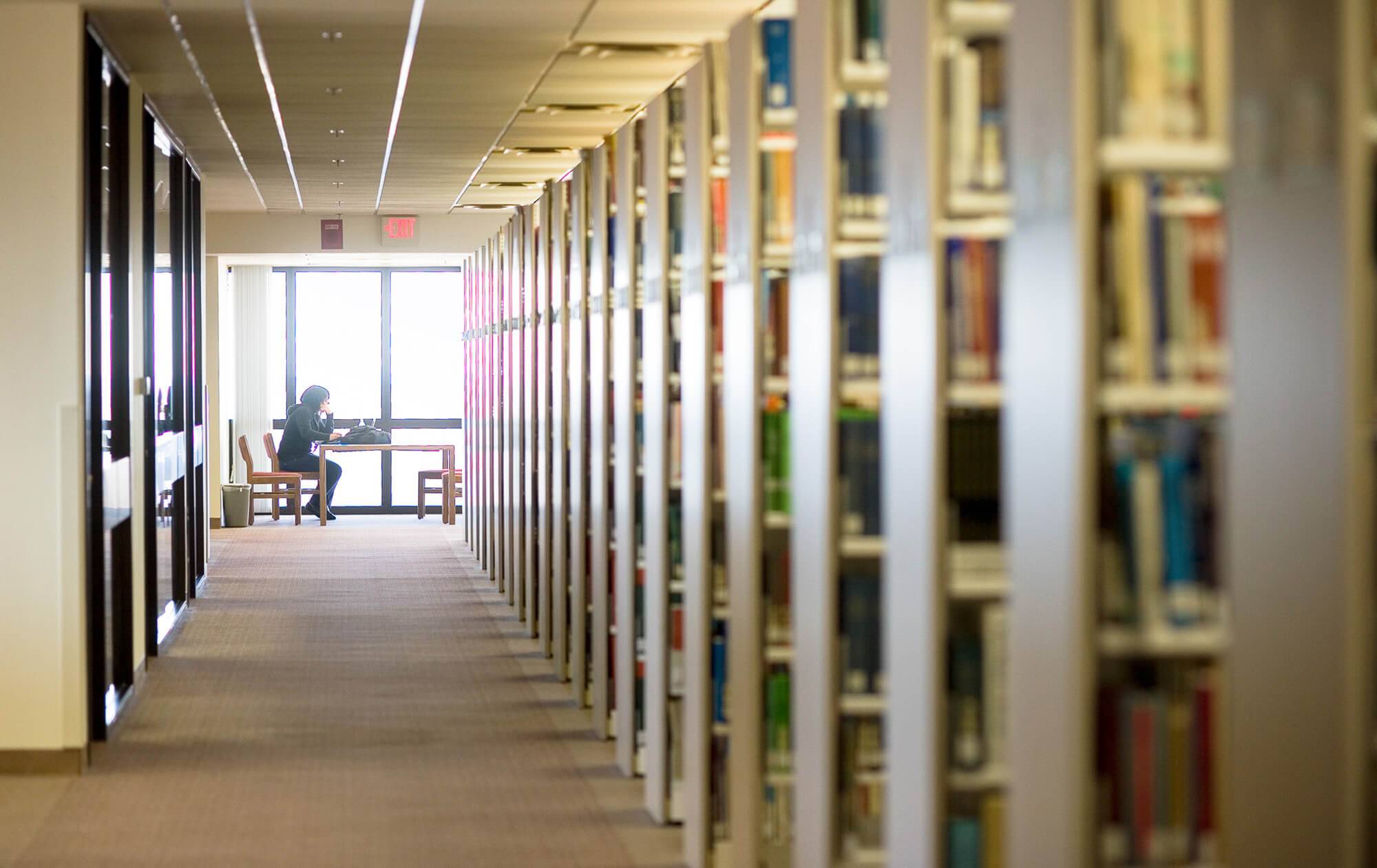 View of the stacks in Alkek ending in a student studying backlit by sun shining through a window