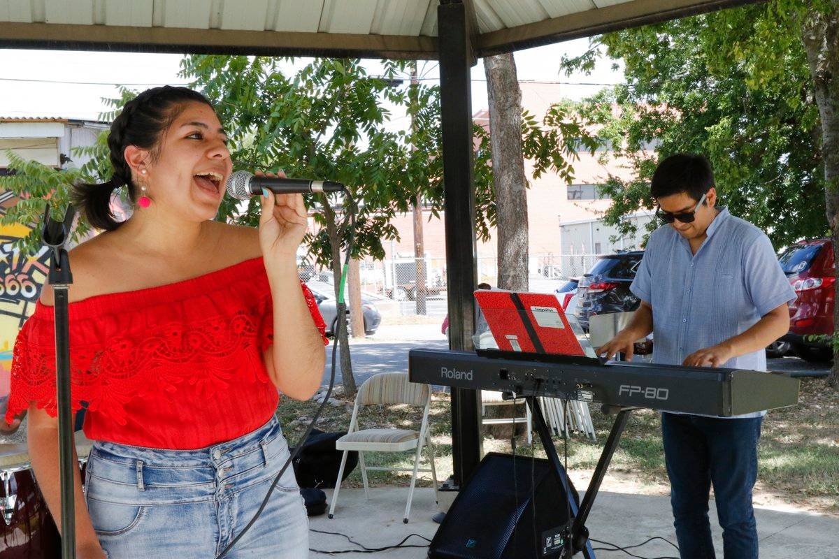 girl singing on the left in a red top wearing jeans, boy playing the keyboard on the right, wearing blue shirt and dark blue jeans and a sunglasses