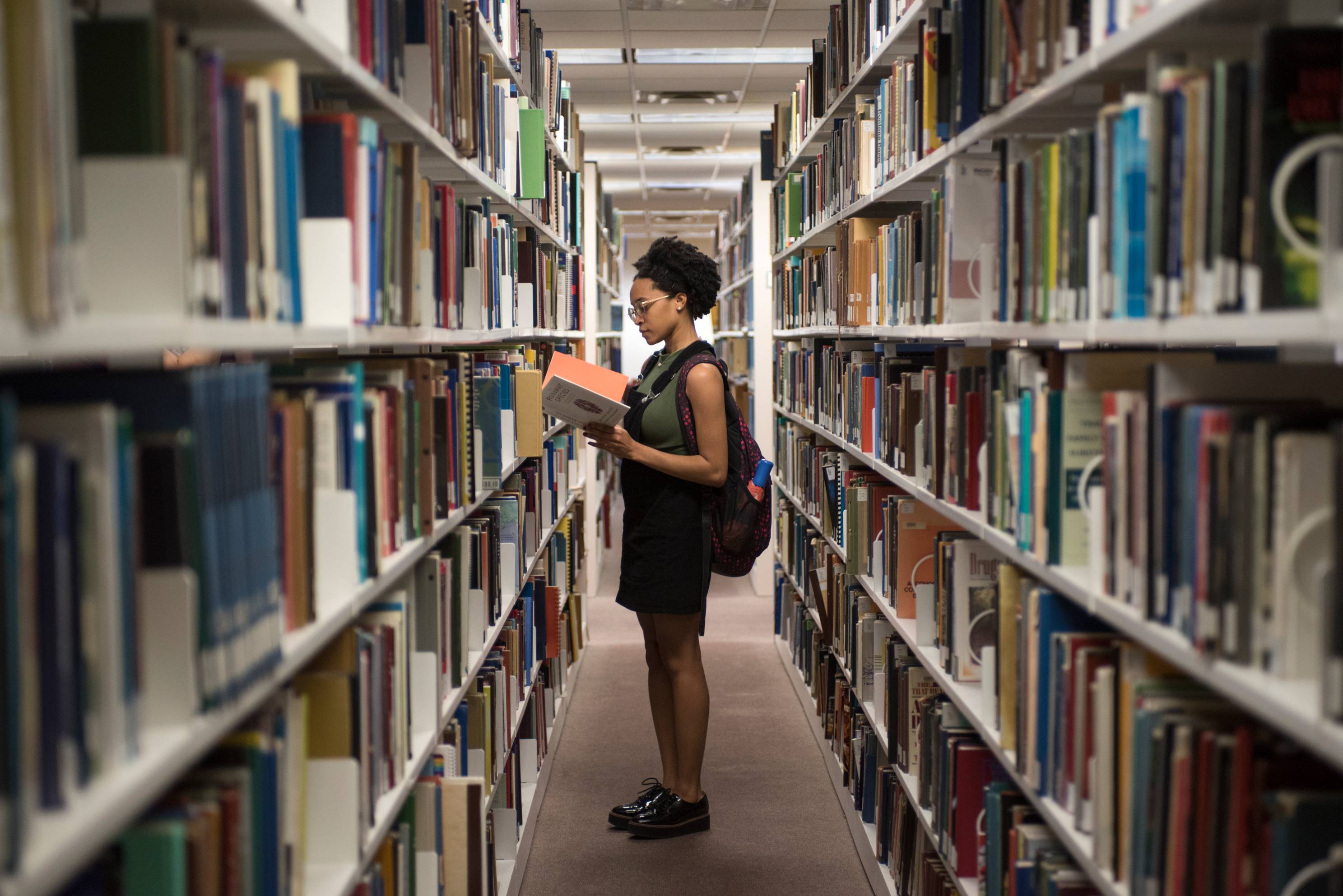 a female student reading a book in the library surrounded by shelves of books