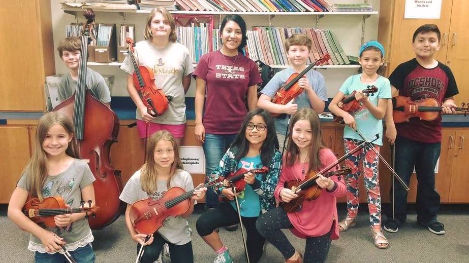 A group of young string students posing with an instructor in a classroom.