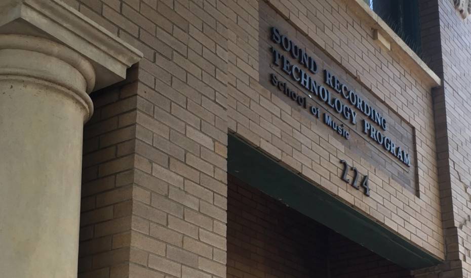 A brown brick building, with a single beige collum visible. A metal sign above an entranceway.