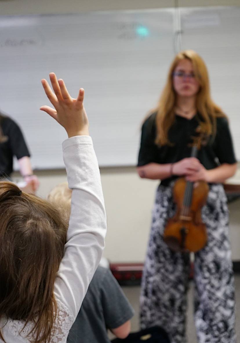 A string teacher stands in front of a classroom as a student raises their hand.