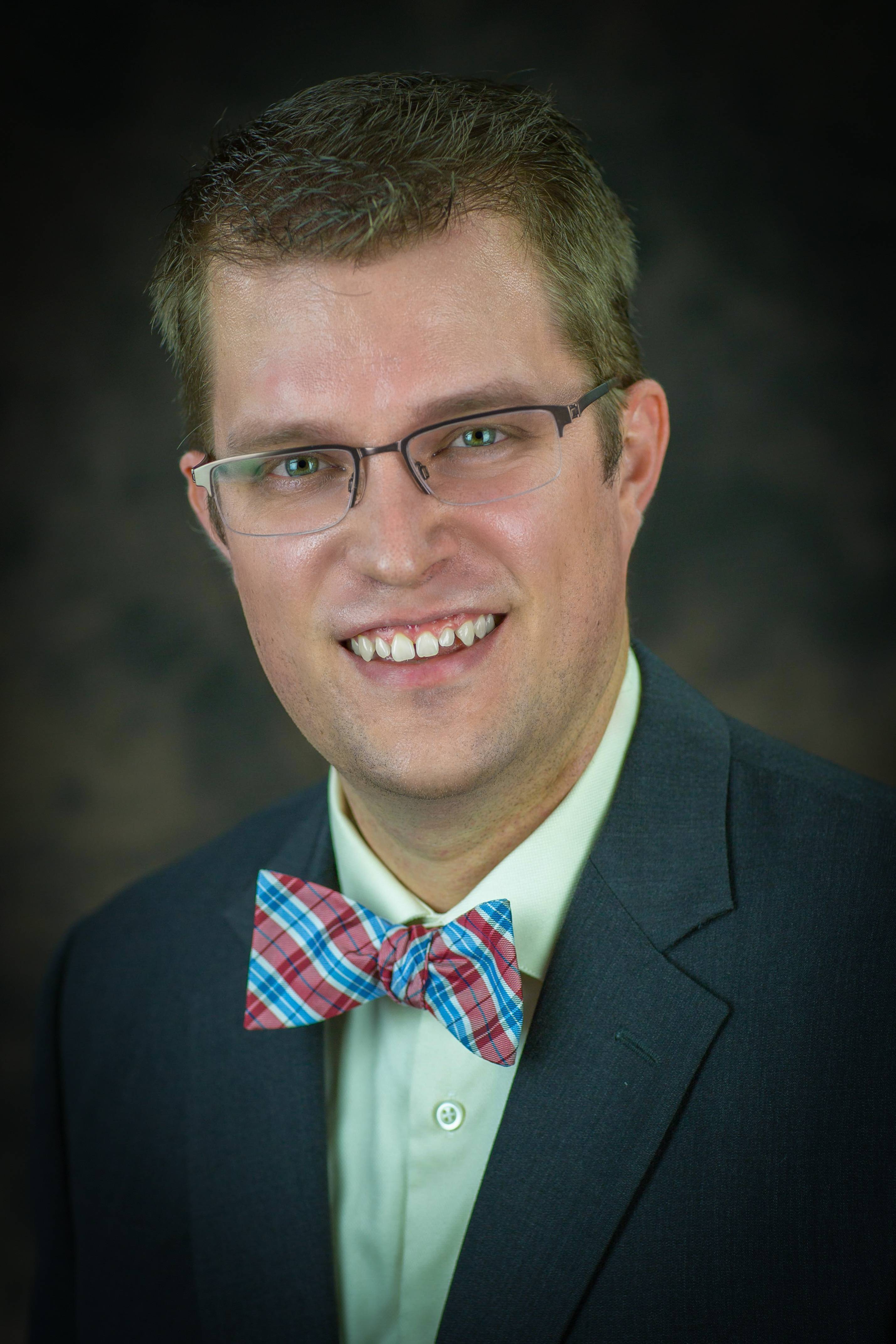 A smiling man wearing a suit jacket with a plaid bow tie.