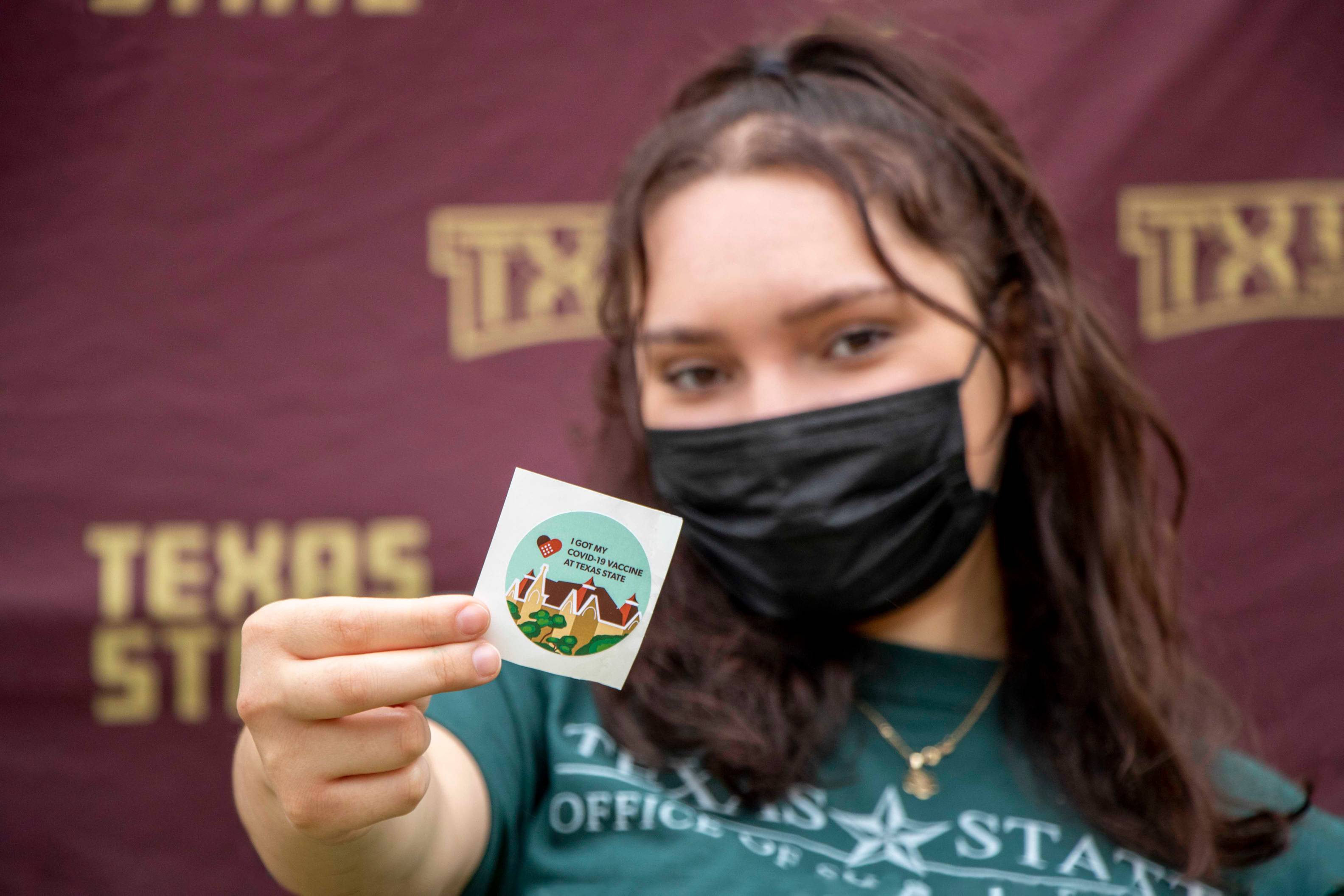 Student with mask holding a sticker for receiving a COVID-19 vaccine.