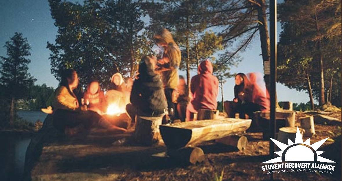 Photo of a group of students speaking with each other while gathered around a campfire.