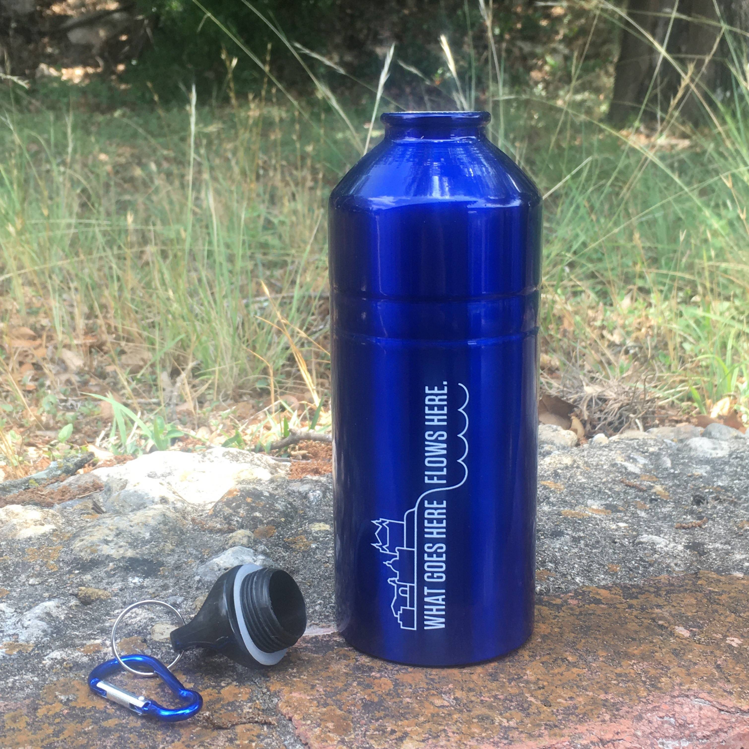 A reusable blue, metal water bottle that says "What Goes Here Flows Here" is sitting on a brick wall with the cap next to it.