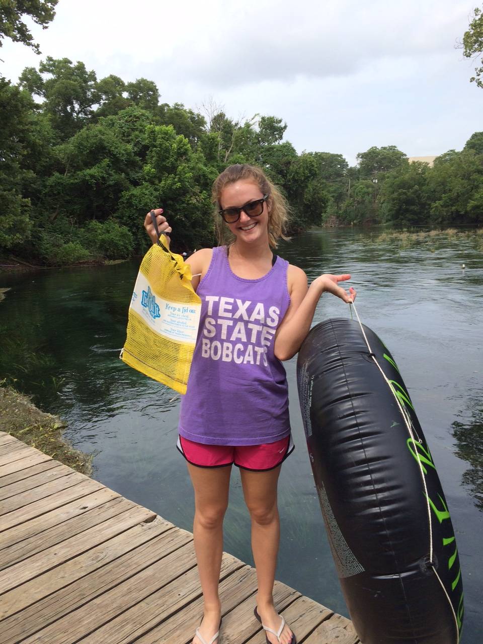 Photo showing a TXST student holding a litter collection bag and a tube on a dock of the San Marcos River.