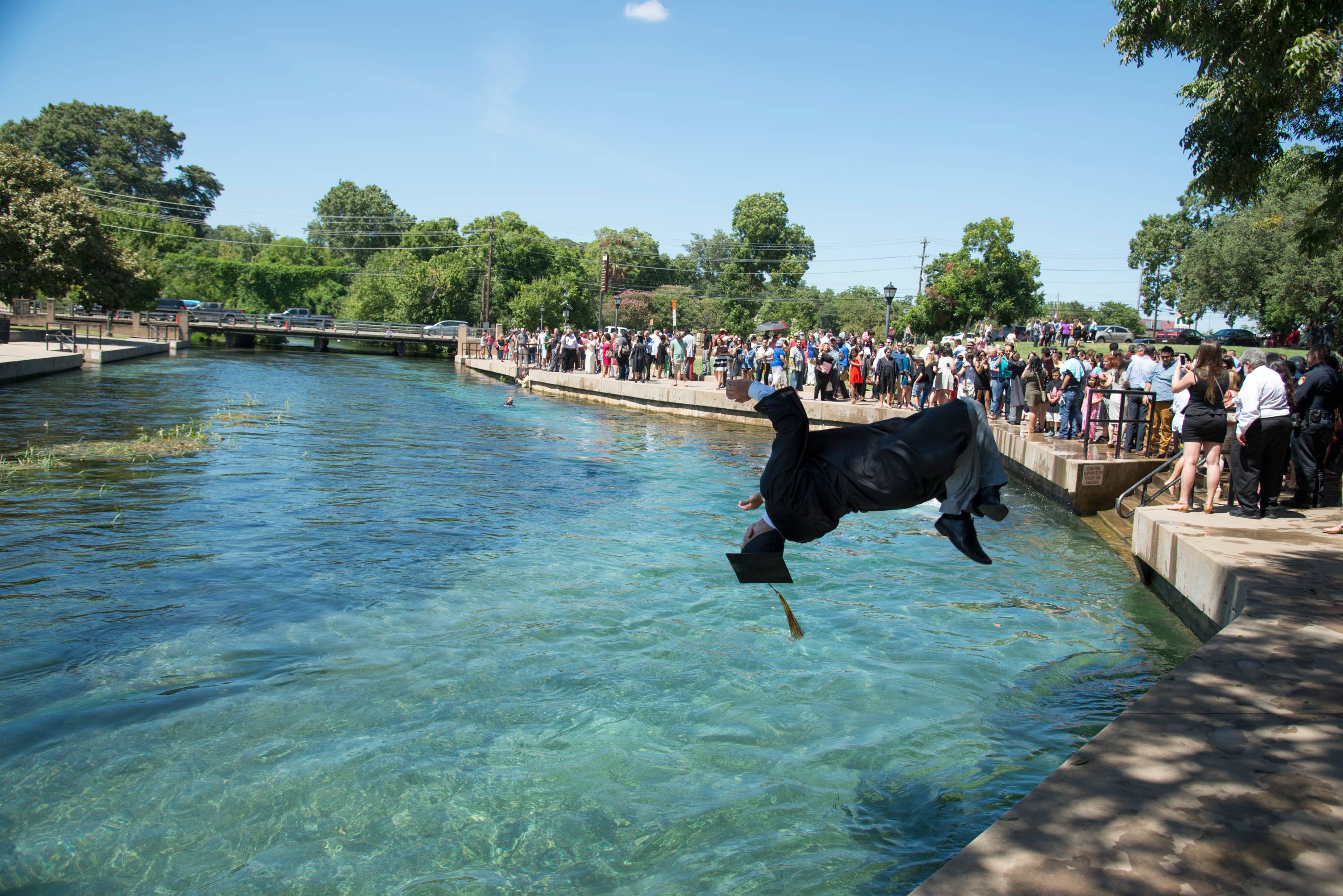 Photo shows a student doing a back flip into the San Marcos River in their graduation cap and gown.