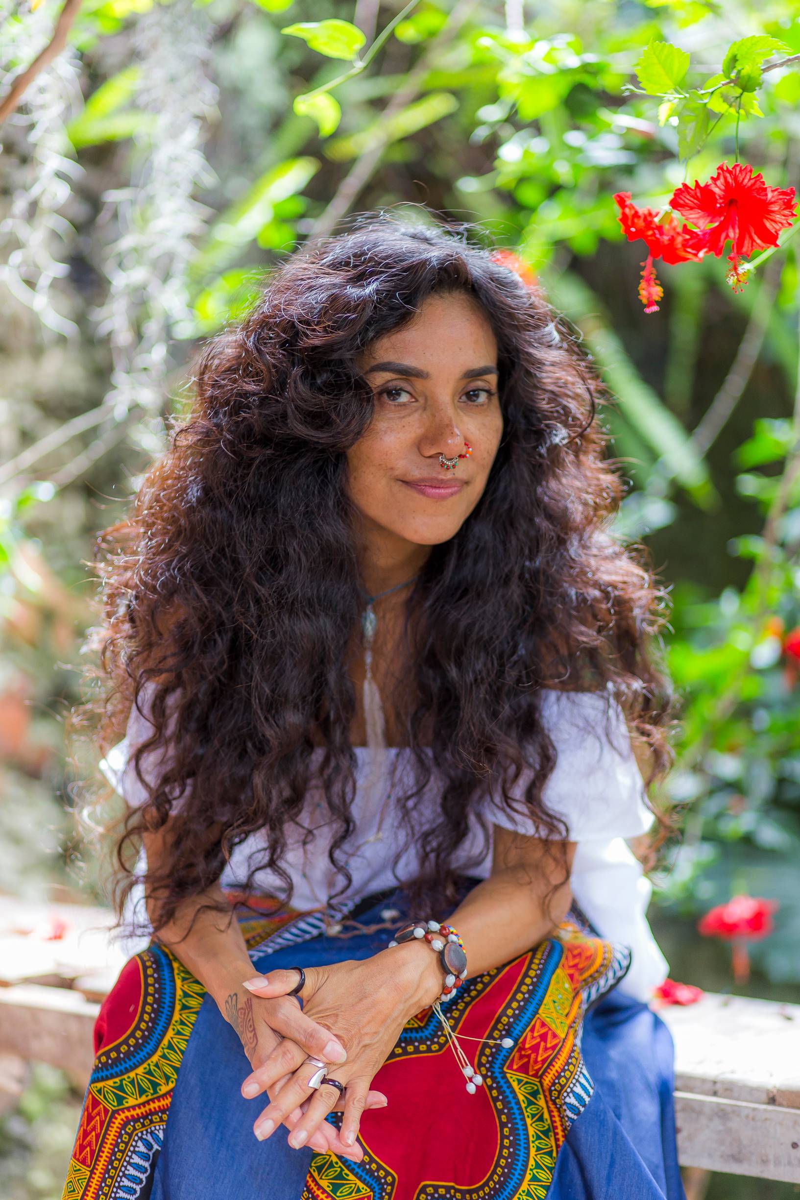 Yuyi Morales sitting on a bench in front of green plants and red flowers