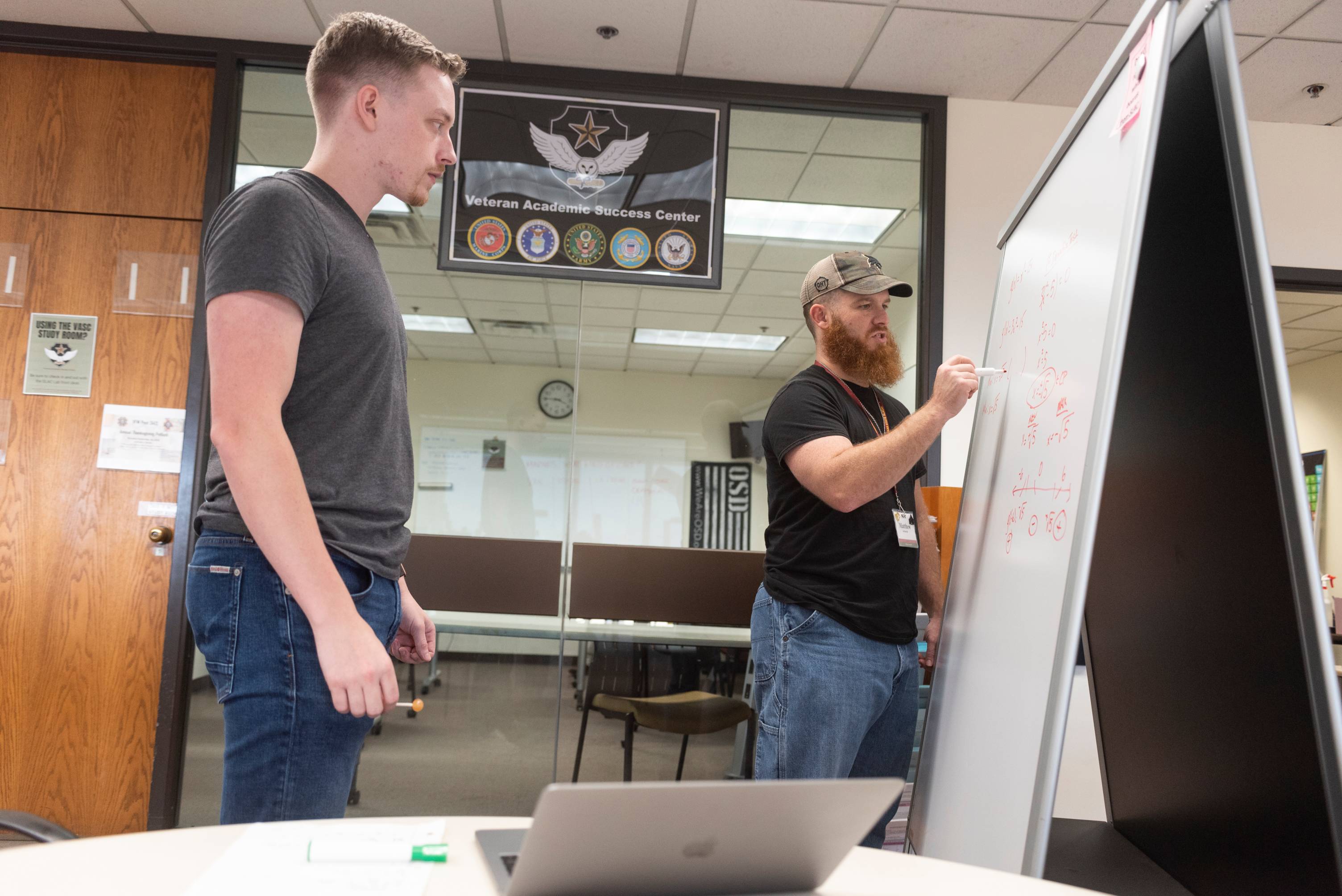 two students stand in front of a sign that says Veteran Academic Success Center. one student is writing on a dry erase board while the other observes.