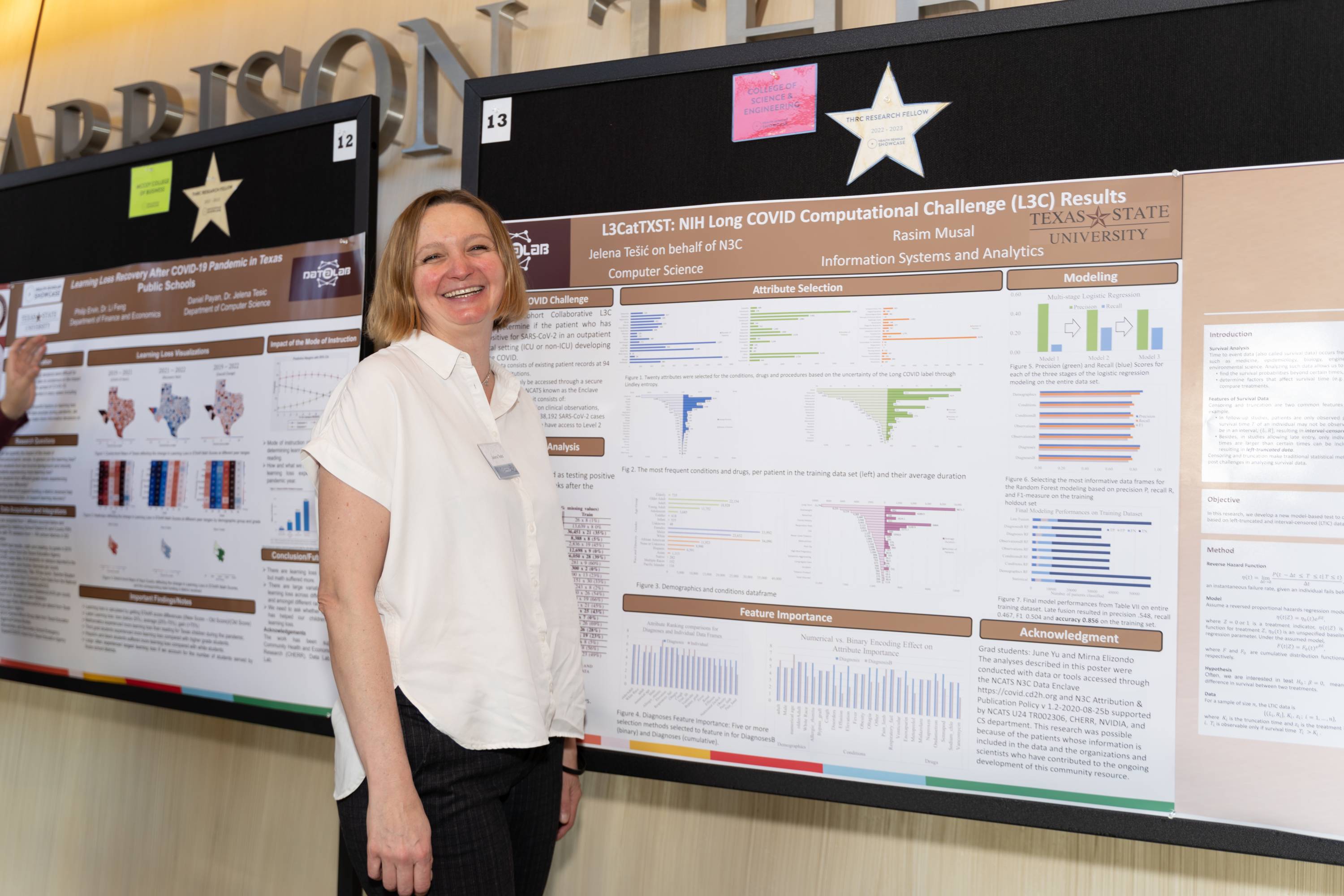 One female research stands on the side of her research poster and smiles into the camera.