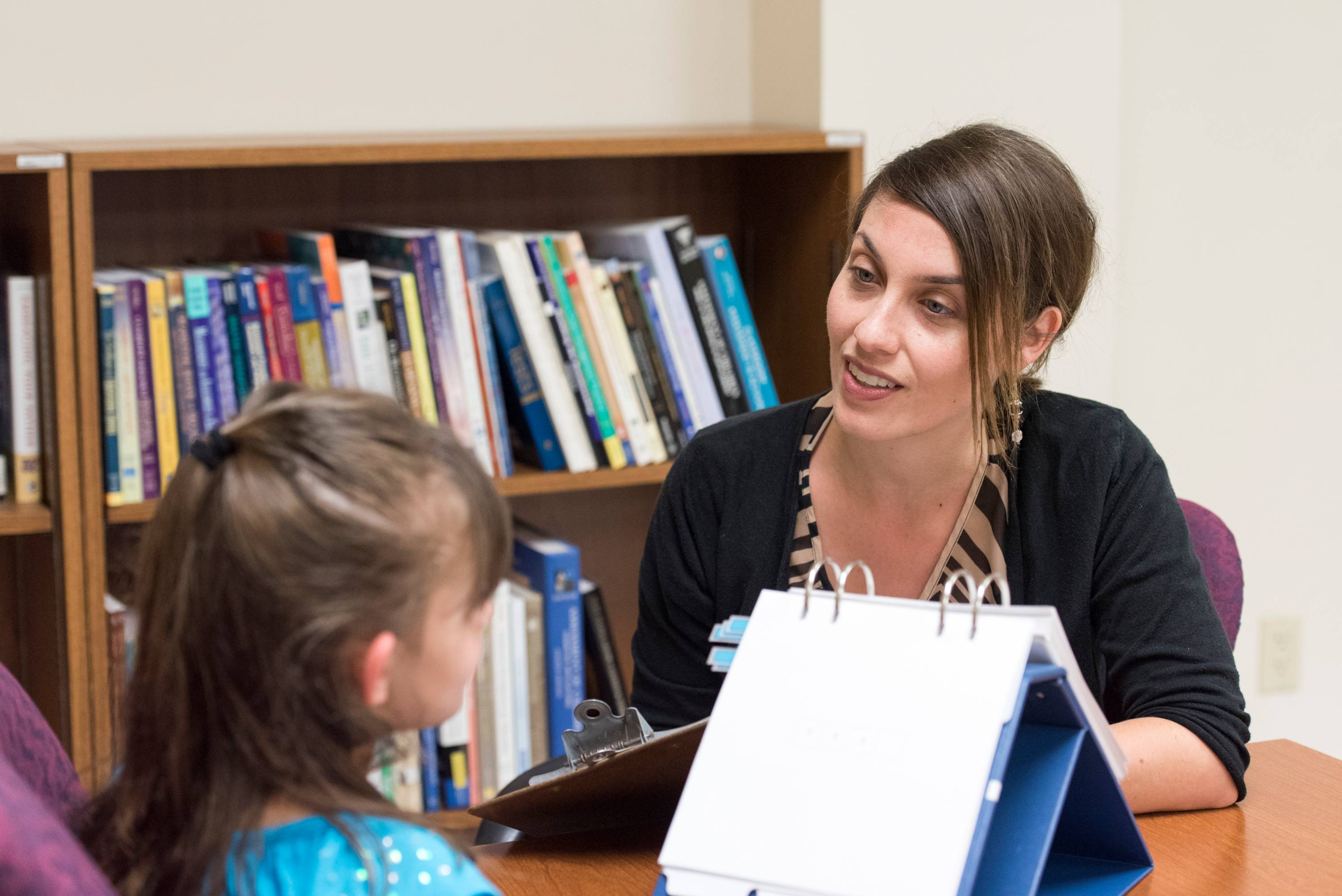 a school psychologists conducts an assessment with a young student, the school psychologist is writing on a clipboard and a small flip chart is situated on the table between the pair